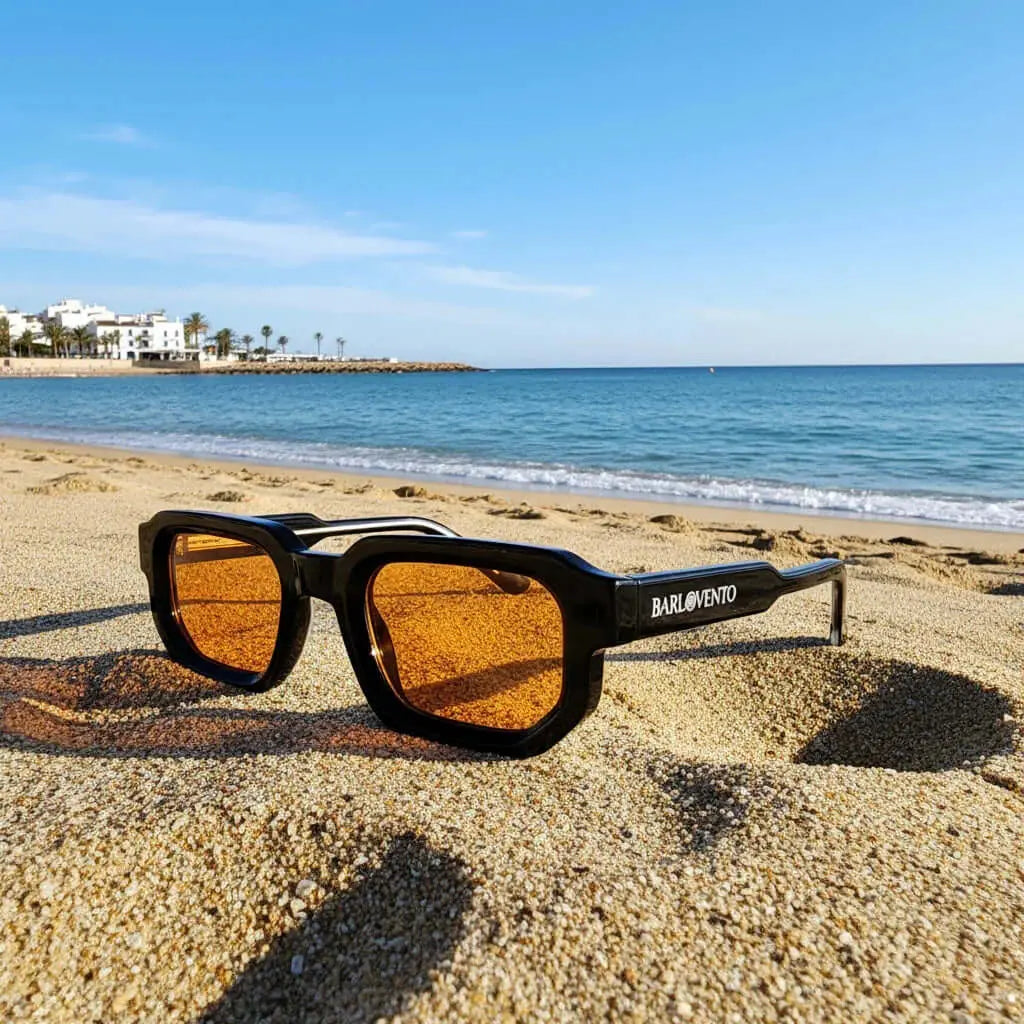 Gafas de sol negras de estilo rectangular con montura gruesa y lentes naranjas sobre la arena de una playa soleada.