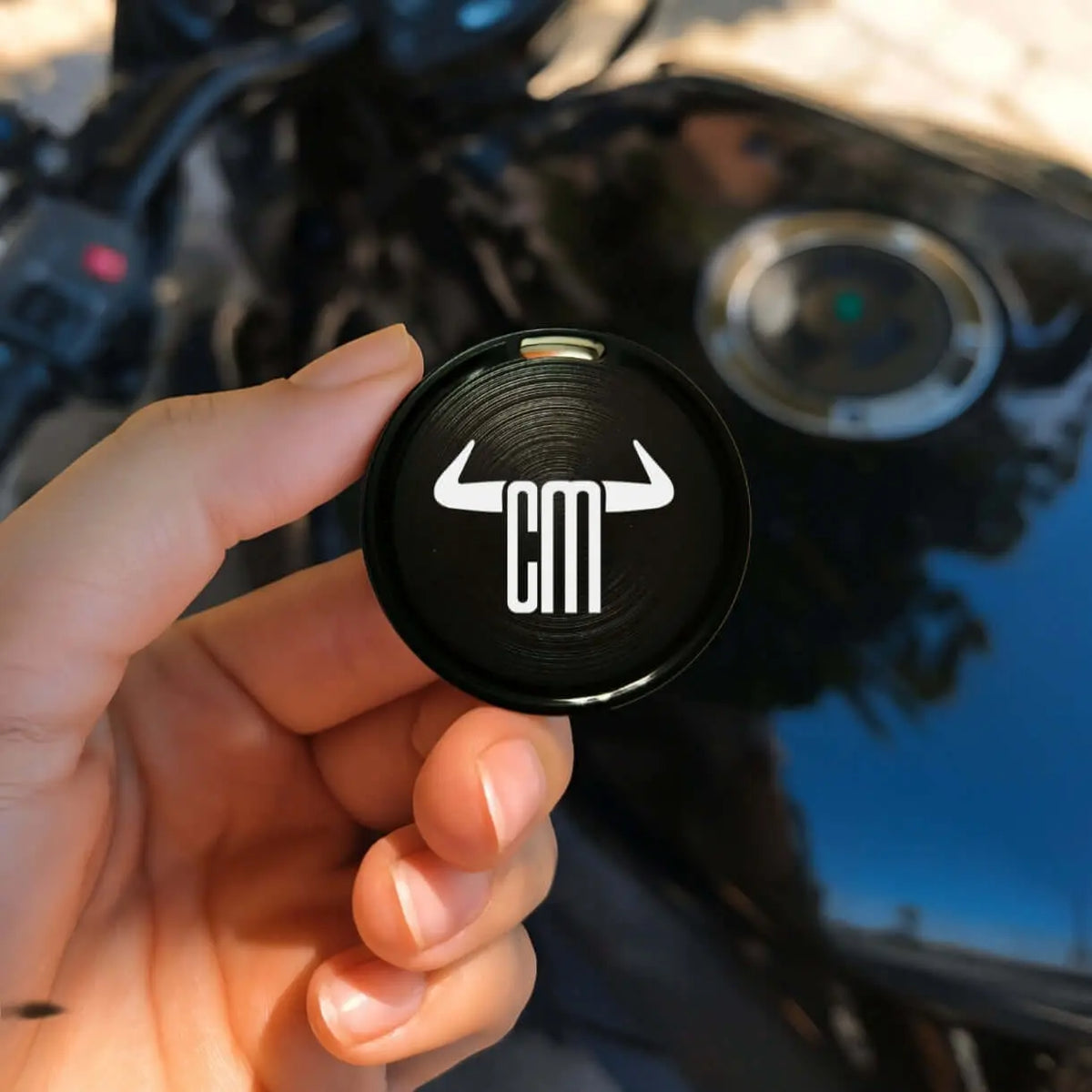 Mano sosteniendo mini localizador GPS negro, circular y metálico con logo blanco ChuchesMoto sobre fondo de motocicleta.