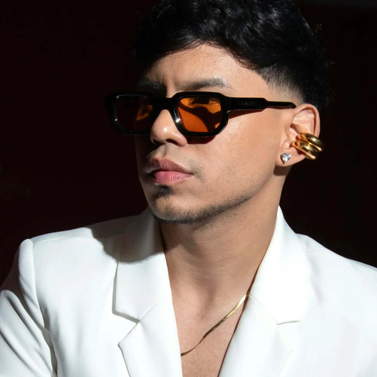 Joven con gafas de sol negras de montura gruesa y lentes naranjas, vistiendo blazer blanco elegante en fondo oscuro.
