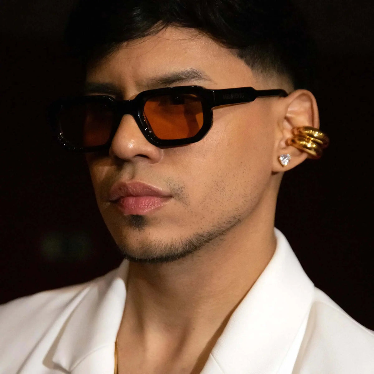 Hombre con gafas de sol rectangulares, montura negra gruesa y lentes naranjas, con chaqueta blanca elegante.