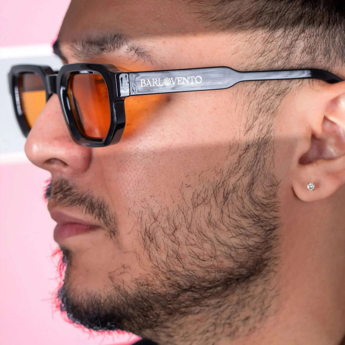 Gafas de sol negras de estilo rectangular con lentes naranjas puestas por un hombre de perfil sobre fondo rosa.