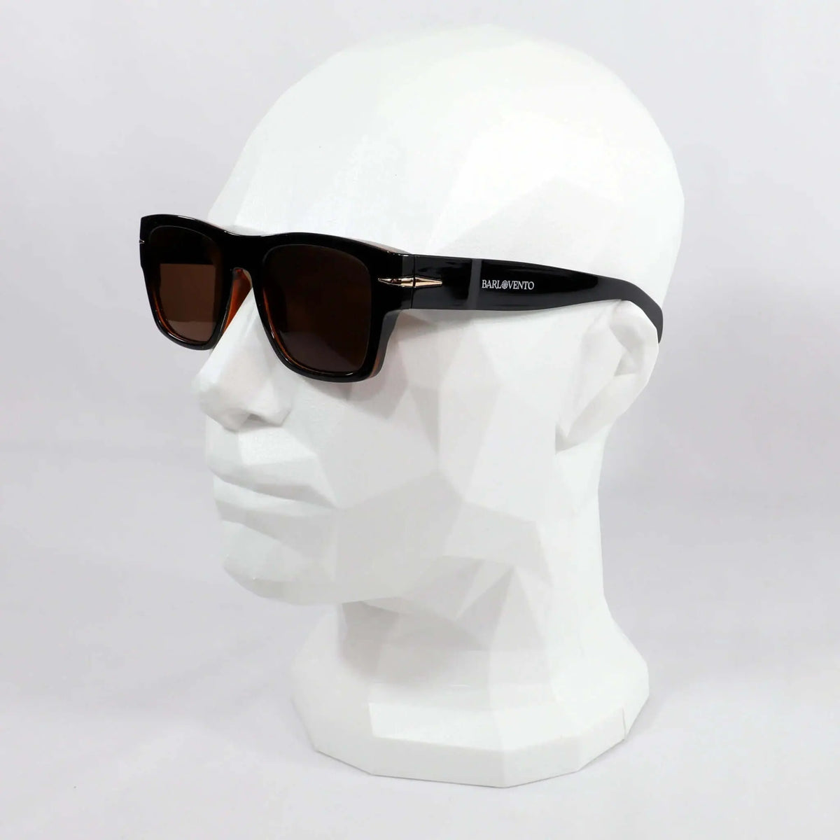 Gafas de sol retro negras con montura rectangular y lentes marrones sobre un busto de exposición blanco.