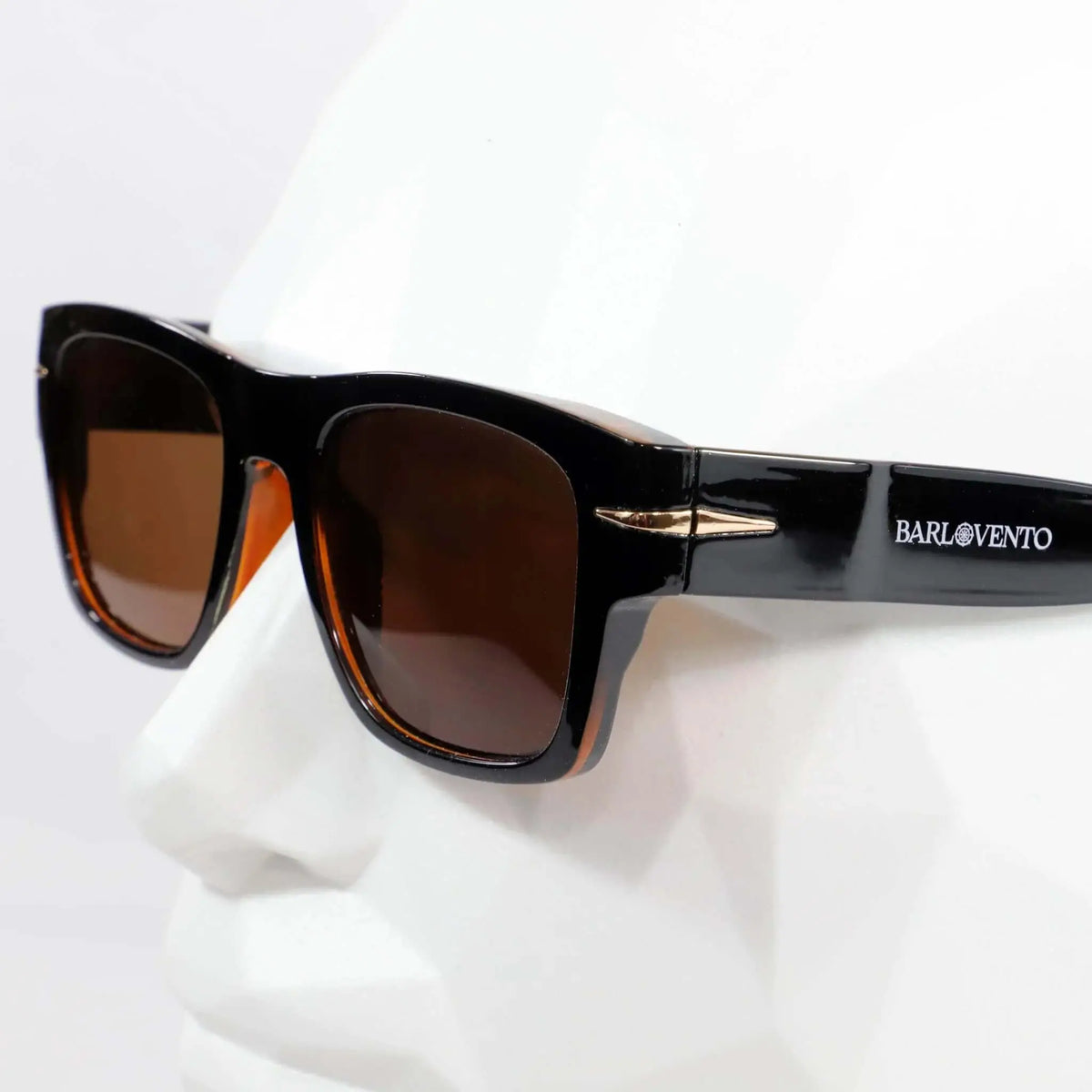 Gafas de sol retro negras con montura cuadrada, acabado brillante y lentes marrones sobre maniquí blanco.
