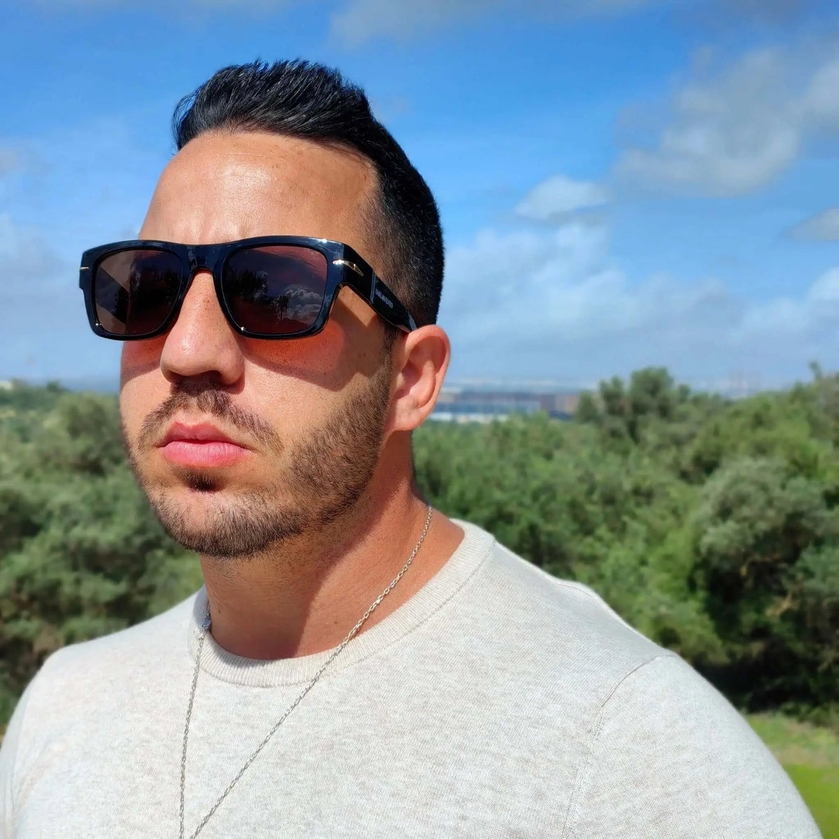 Hombre con gafas de sol negras retro, montura gruesa y cristales marrones, con jersey beige en un entorno natural.
