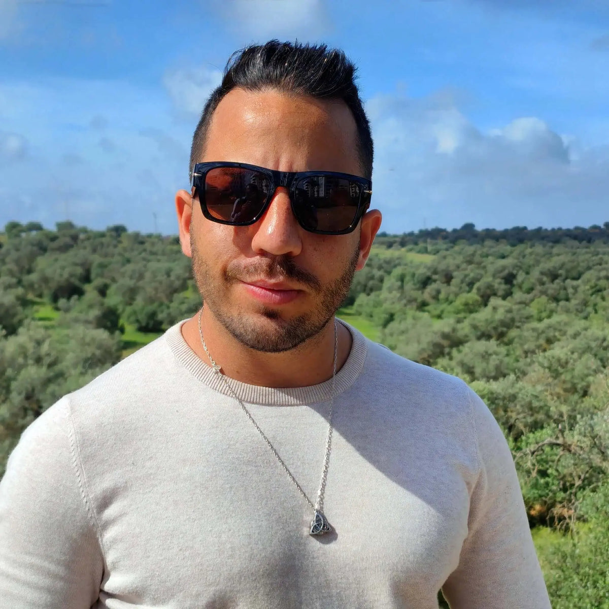 Hombre con jersey beige y gafas de sol retro de montura negra y lentes marrones al aire libre.
