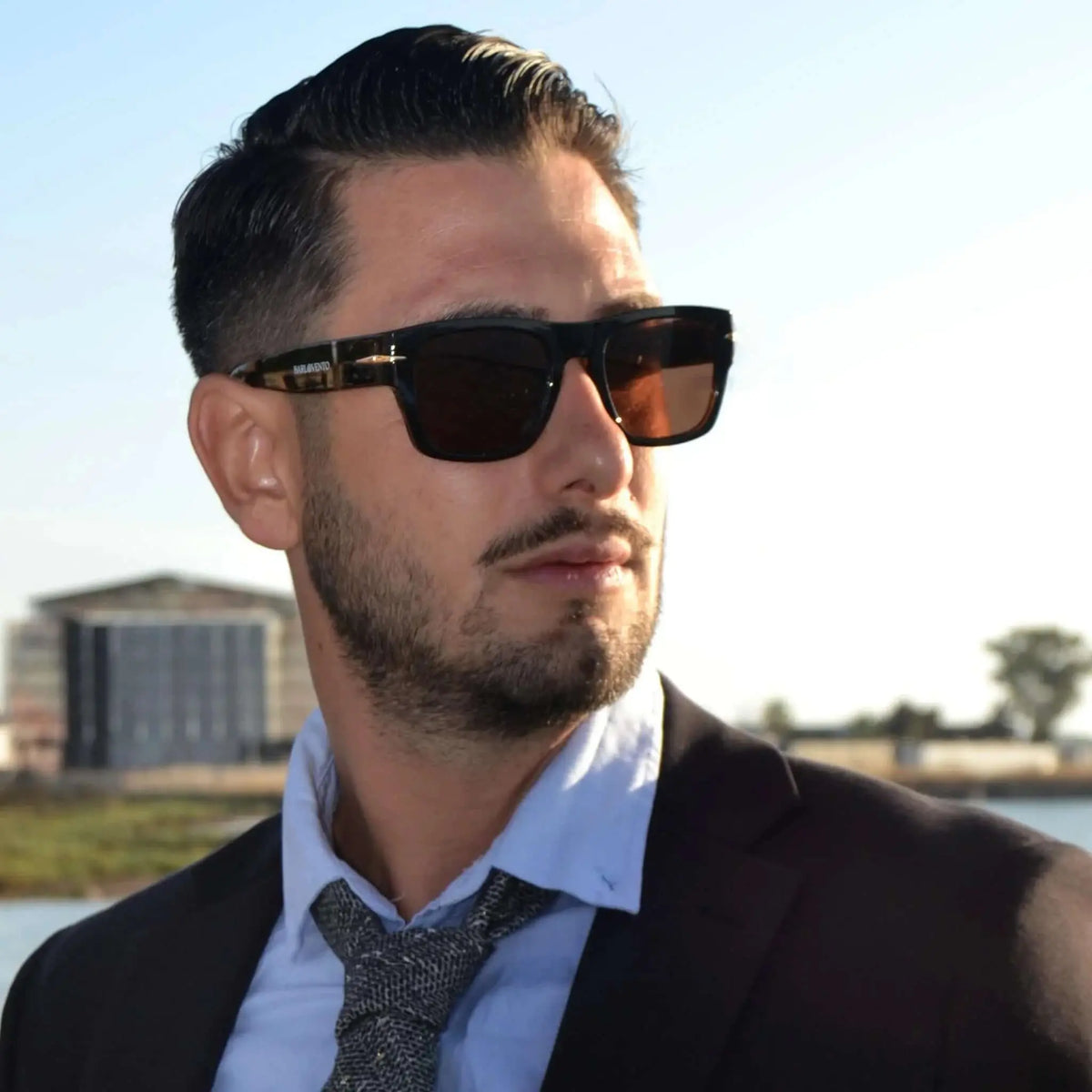 Hombre de traje con gafas de sol retro de montura negra y lentes marrones en exteriores. Estilo elegante y profesional.