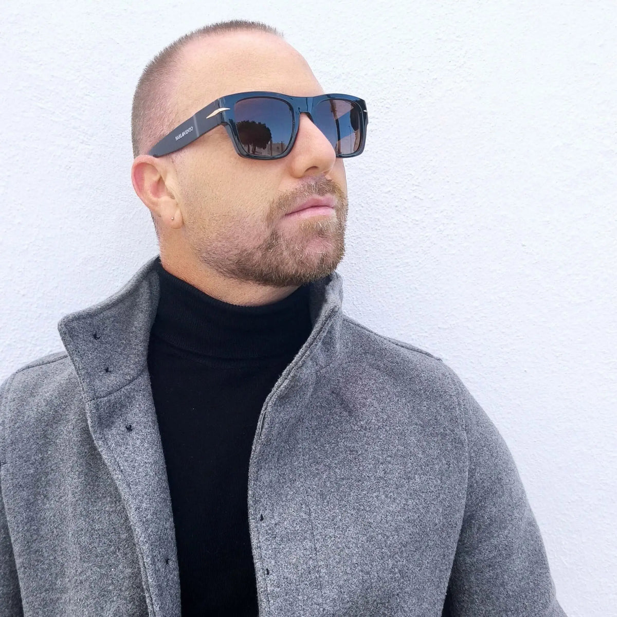 Hombre con barba usa gafas de sol retro negras con lentes marrones, cuello alto negro y abrigo gris de lana.