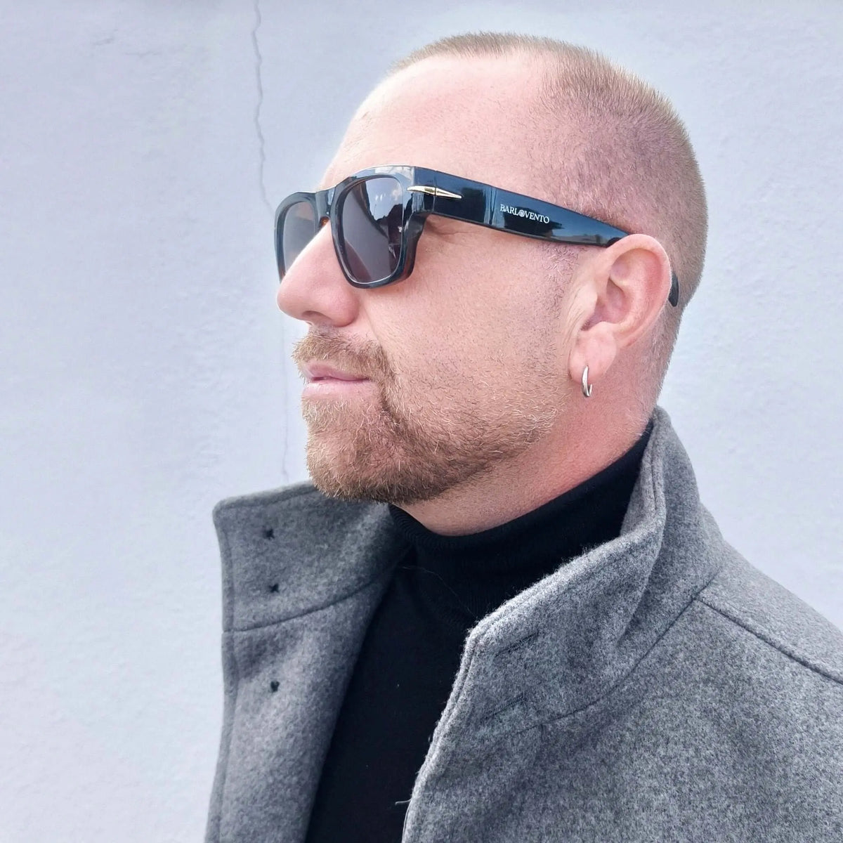 Hombre con gafas de sol retro negras, montura de acetato brillante, lentes marrones y abrigo gris de lana.