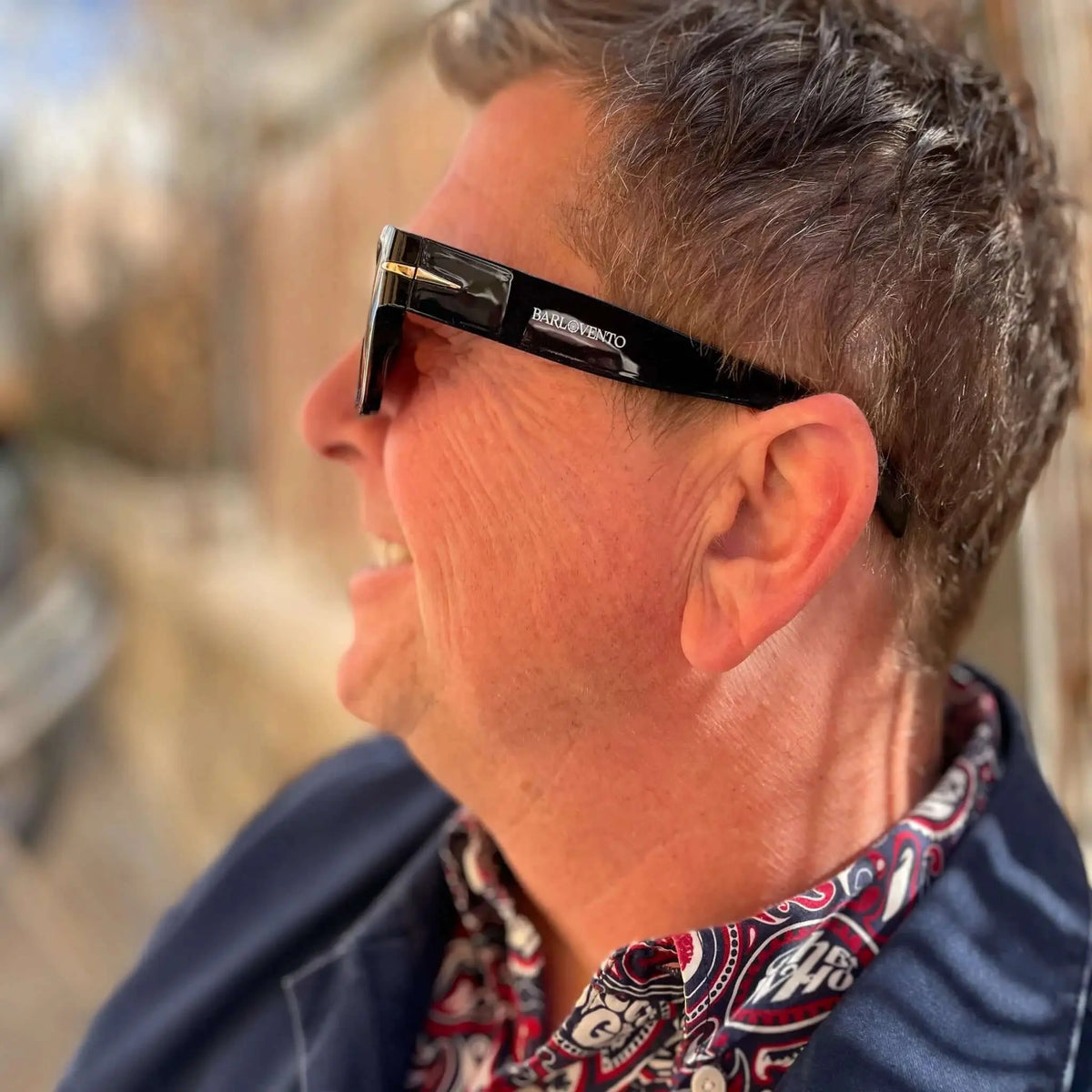 Persona de perfil con gafas de sol negras de estilo retro y acabado brillante, con lentes marrones y detalles dorados.
