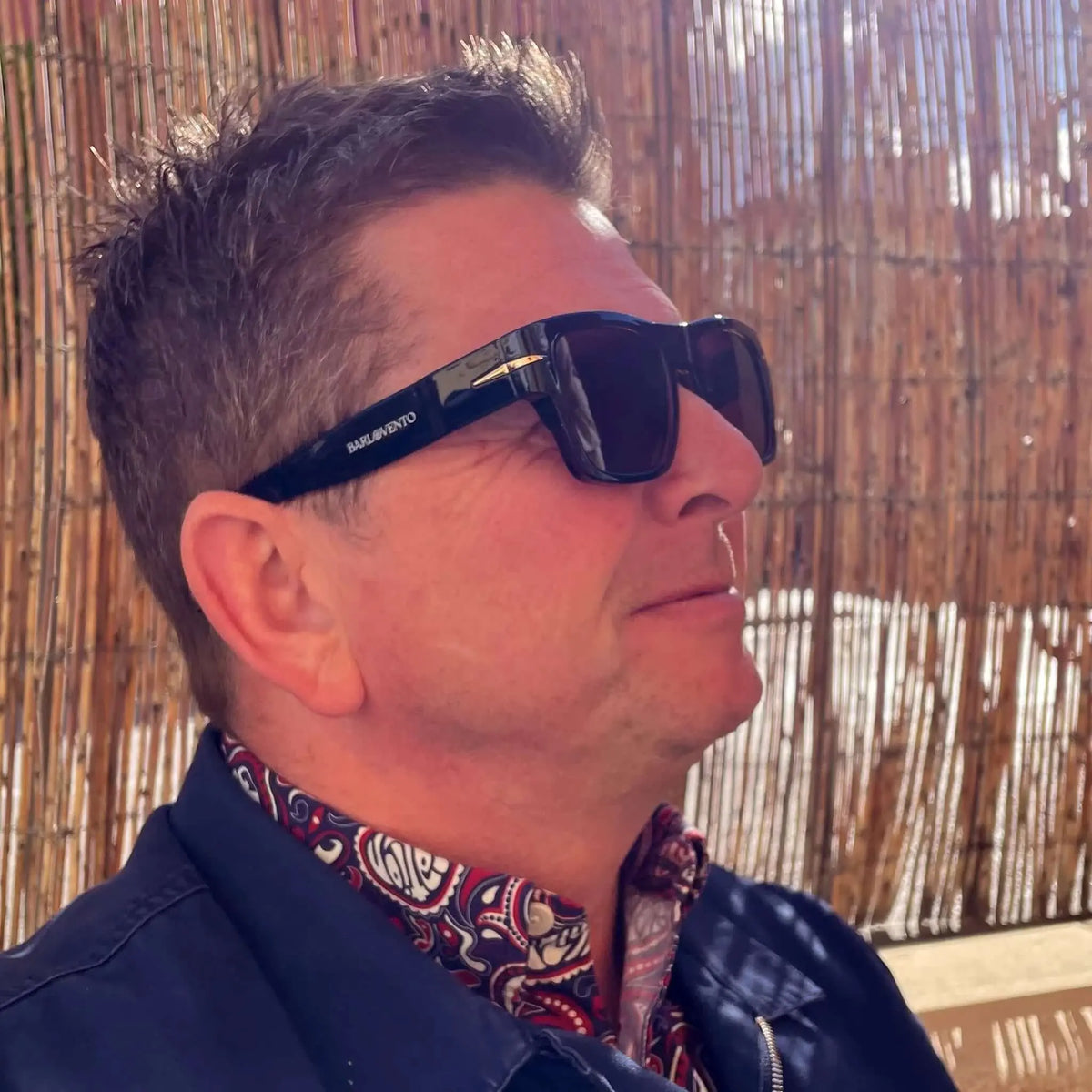 Hombre con gafas de sol negras retro, montura gruesa y lentes marrones oscuros, vistiendo camisa estampada y chaqueta.