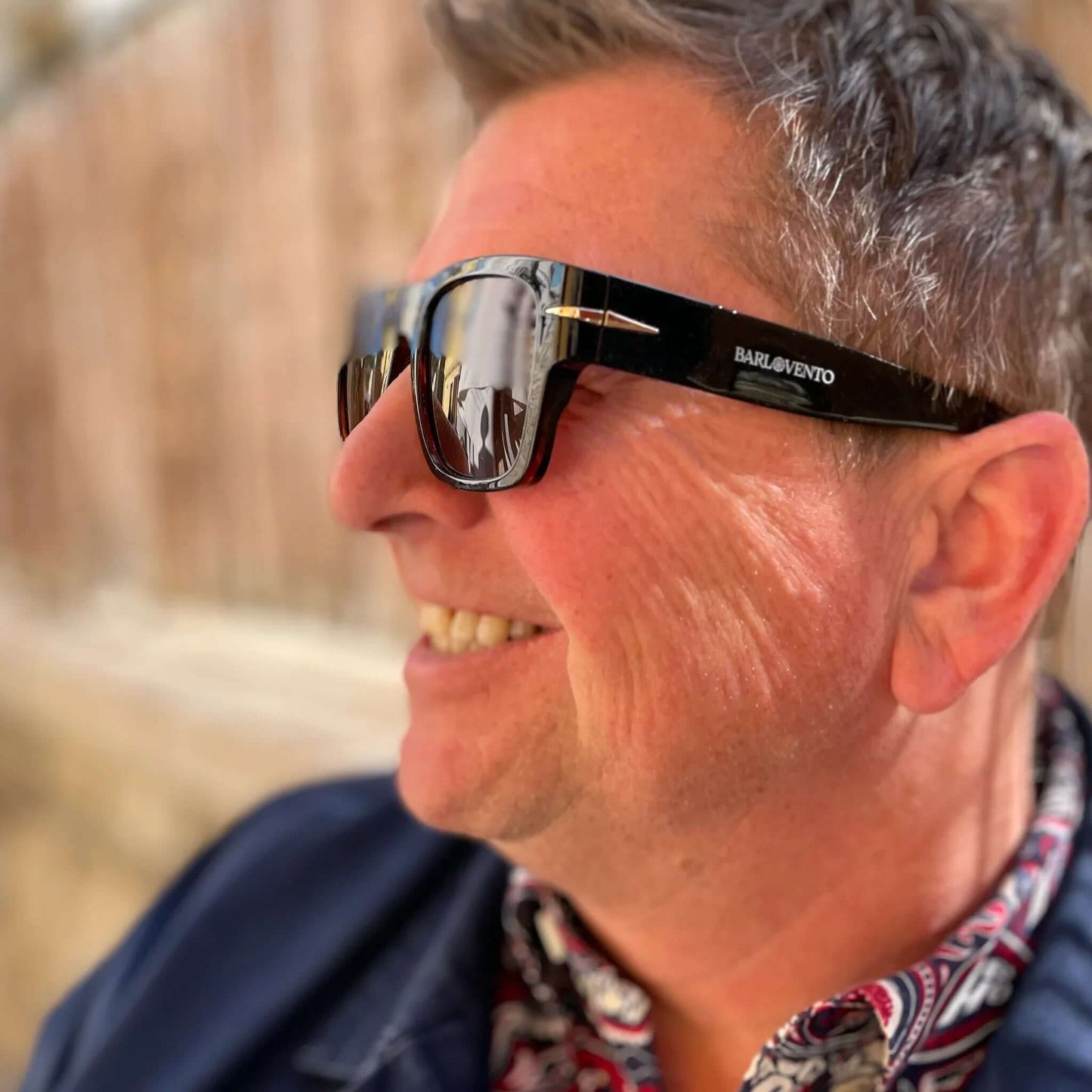 Hombre sonriente con gafas de sol retro negras, montura de acetato brillante y lentes marrones de estilo premium.