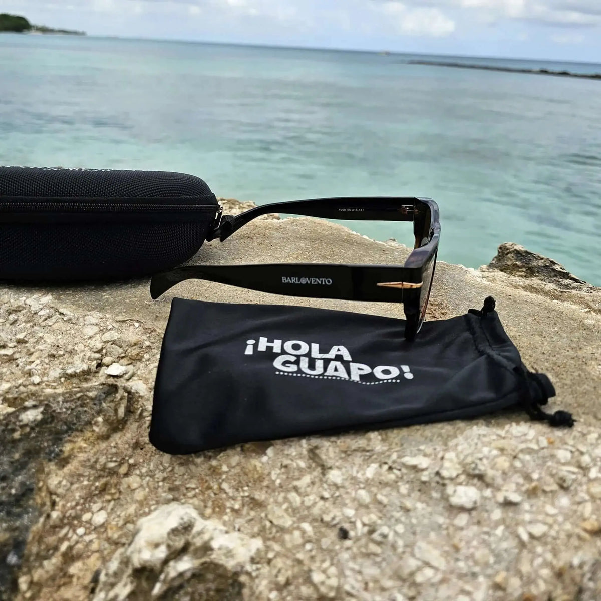 Gafas de sol negras retro con lentes marrones y funda sobre roca frente al mar. Estilo premium, elegante y veraniego.