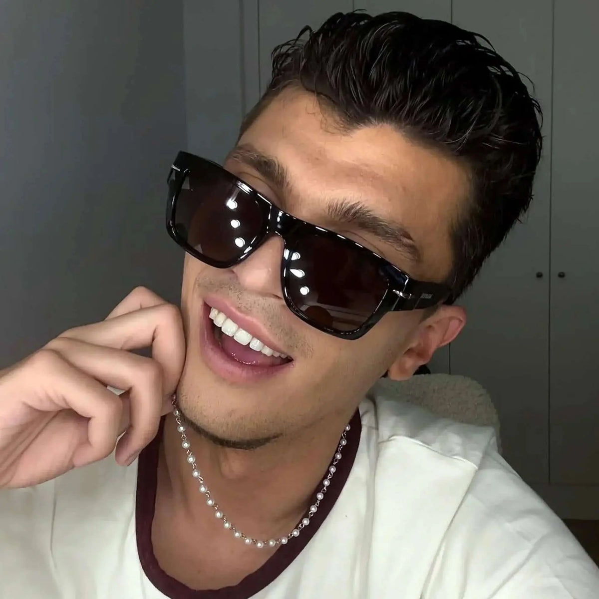 Joven sonriente con gafas de sol retro de montura negra y cristales marrones, estilo elegante con collar de perlas.