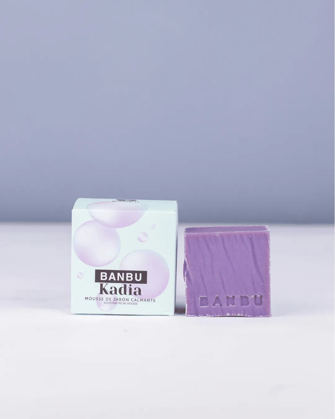 Mousse facial ecológico Kadia de Banbu: pastilla morada junto a su caja con burbujas sobre fondo azul suave.