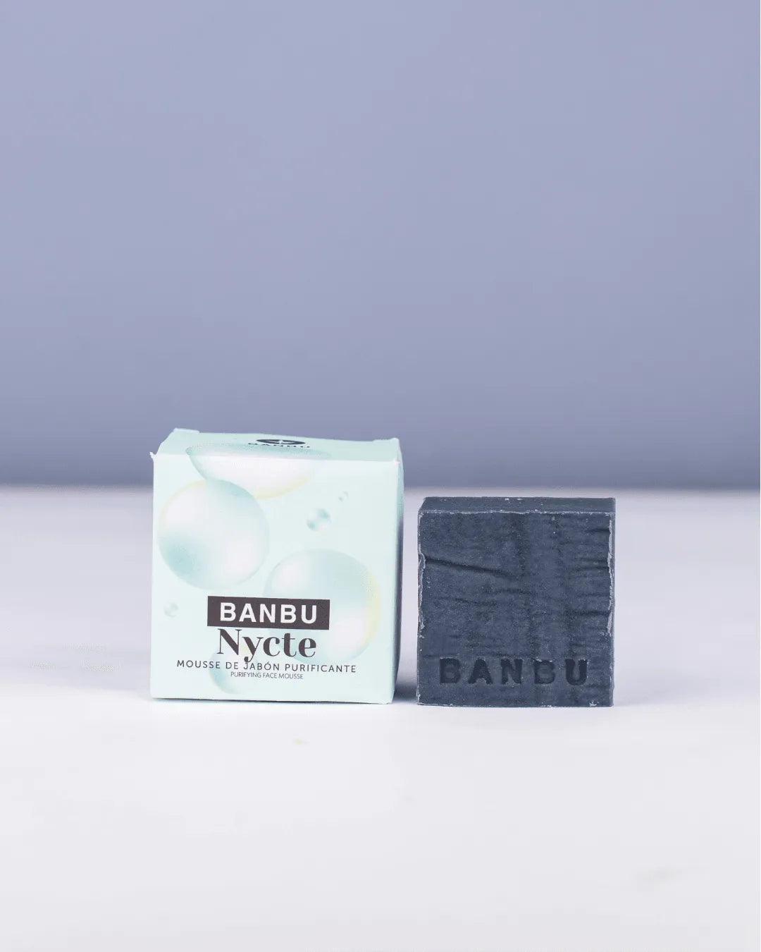 Pastilla de jabón gris oscuro Banbu junto a su caja de cartón verde claro con burbujas, sobre fondo gris suave.