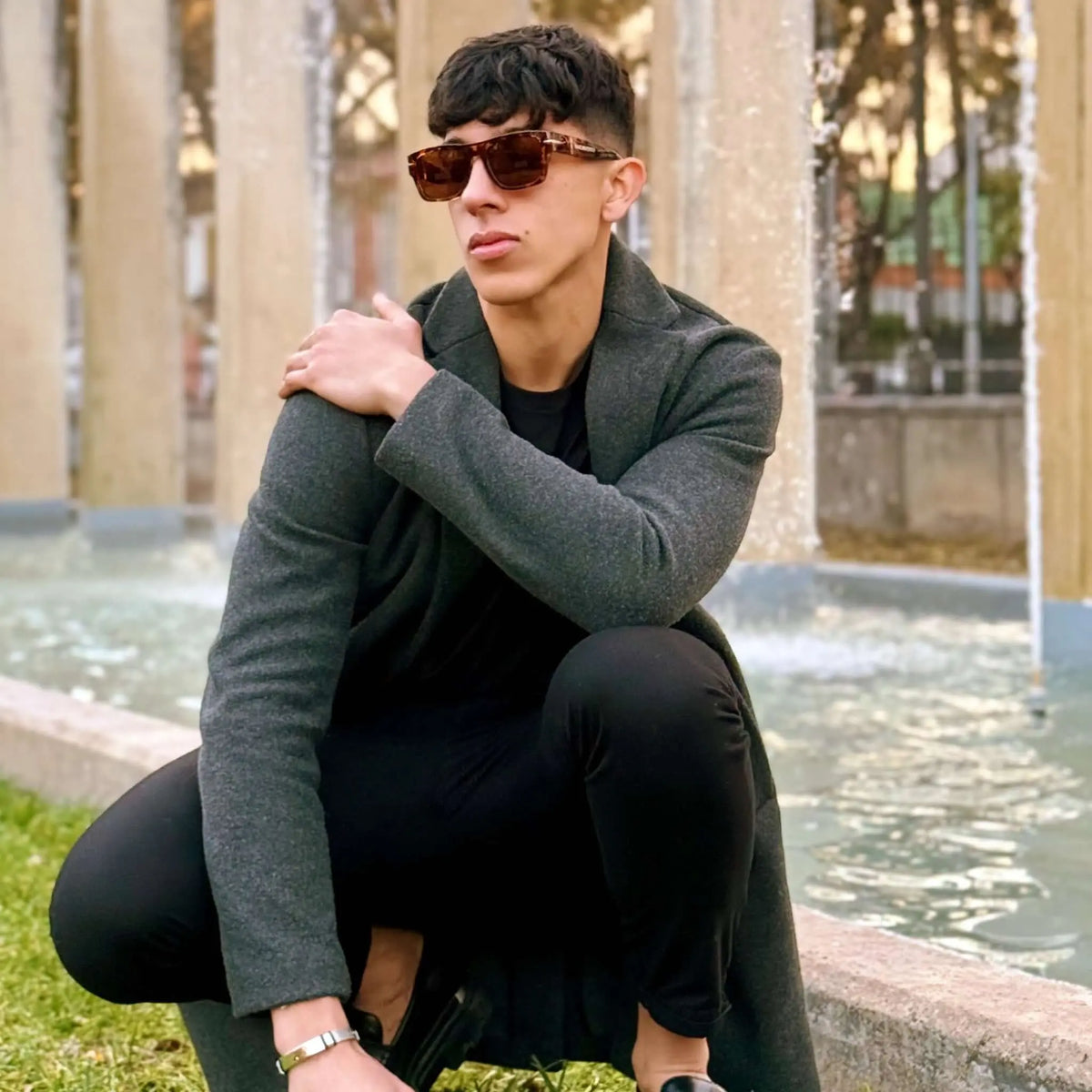 Hombre con gafas de sol retro rojas de pasta, estilo urbano elegante, posando frente a una fuente en el exterior.