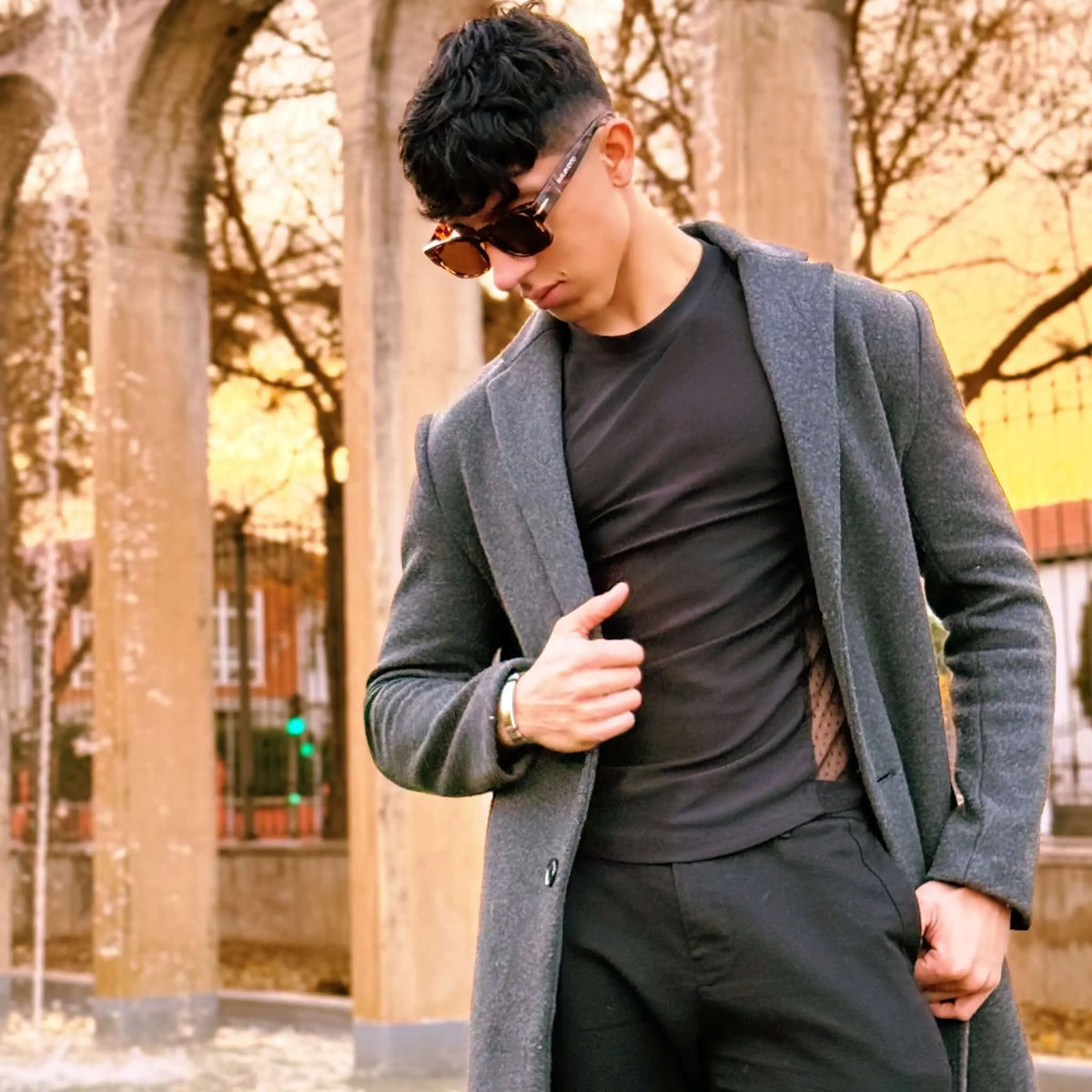 Joven con abrigo gris y gafas de sol retro de montura roja oscura con acabado brillante y estilo elegante.