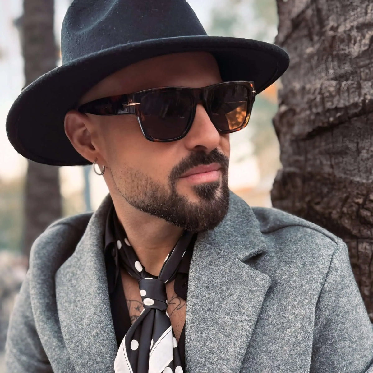 Hombre con gafas de sol retro estilo carey, sombrero negro y abrigo gris, estilo elegante y moderno.