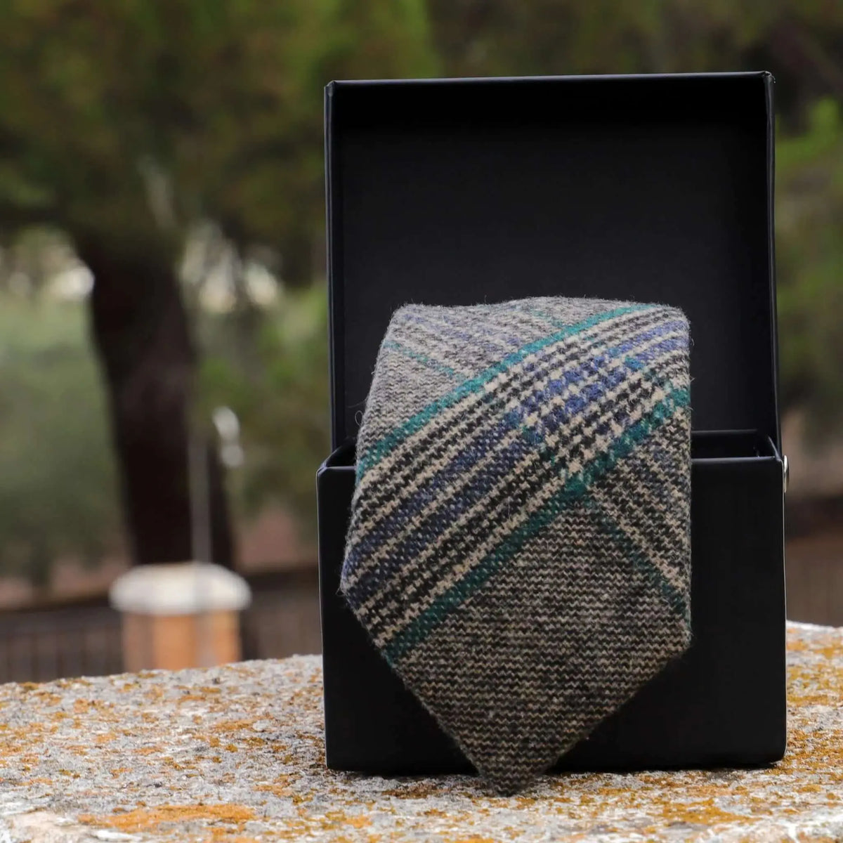 Corbata de lana gris a cuadros con rayas azules y verdes en caja negra sobre superficie de piedra. Estilo formal cálido.