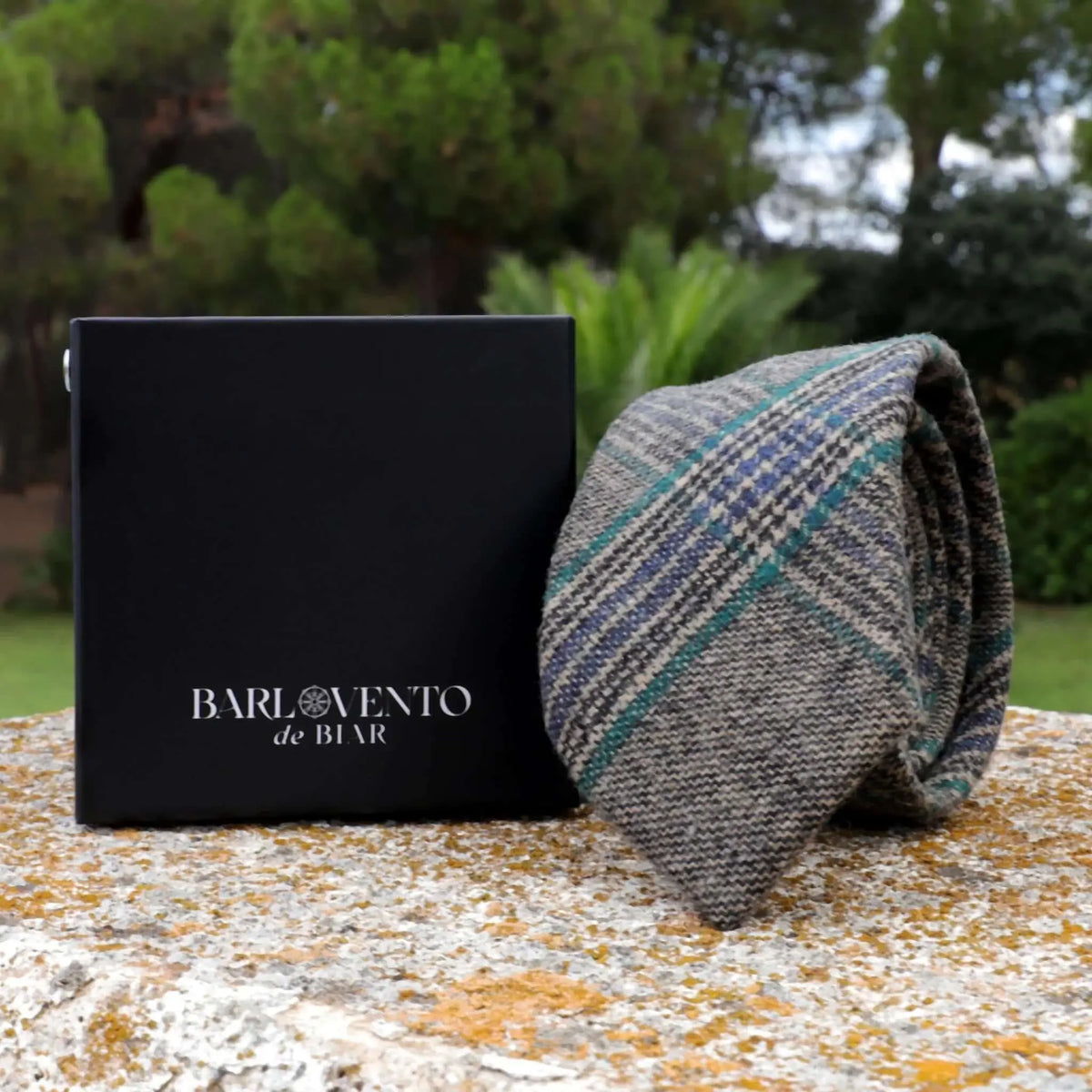 Corbata de lana gris con cuadros azules y verdes, textura tejida, junto a una caja negra en un entorno exterior.
