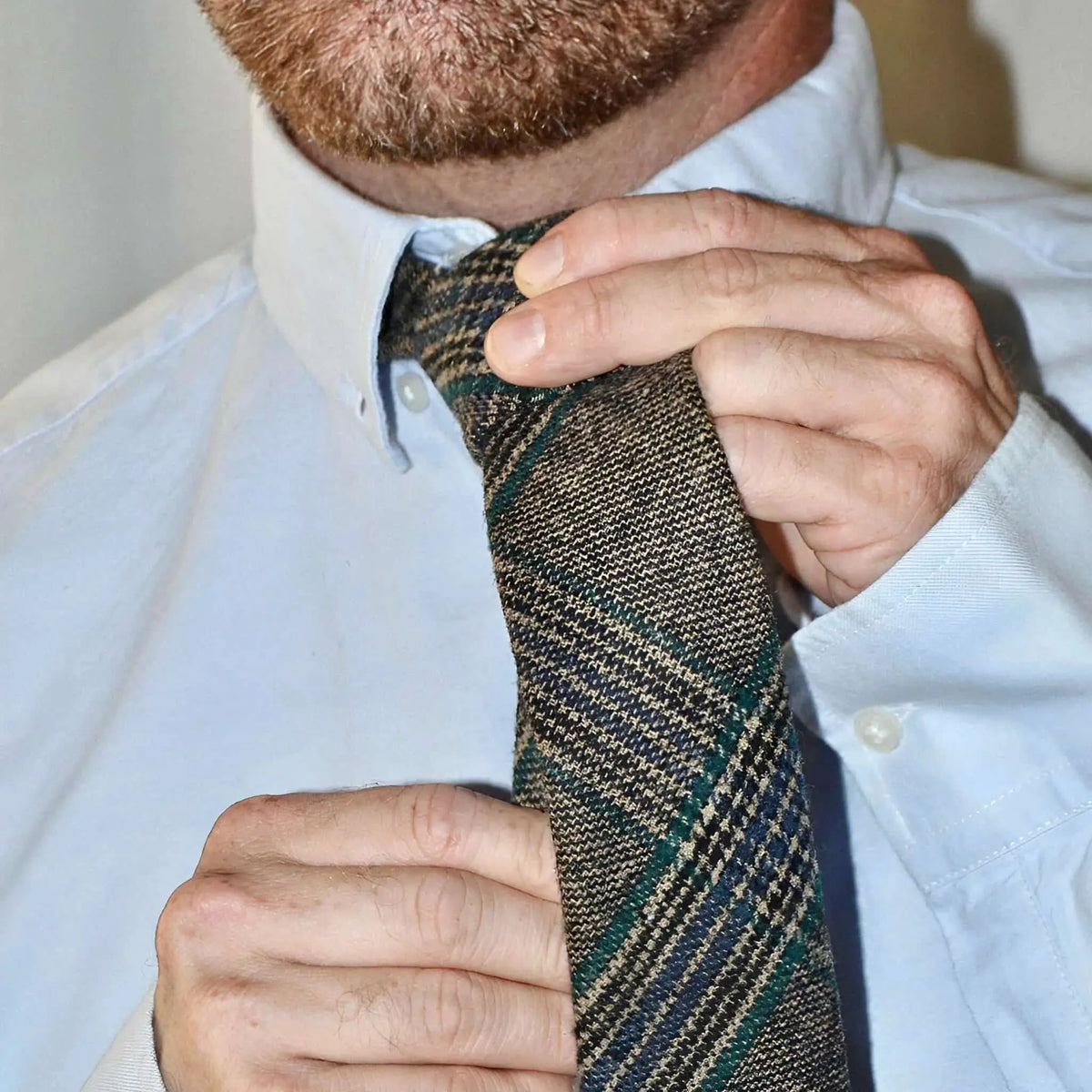 Hombre ajustando una corbata de lana con patrón de cuadros en tonos tierra, verde y azul sobre una camisa azul claro.