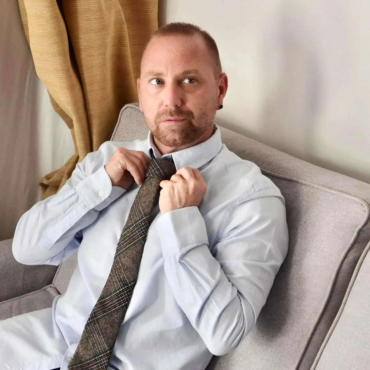 Hombre con camisa azul ajustándose una corbata de lana a cuadros marrones con textura, sentado en un sofá gris.