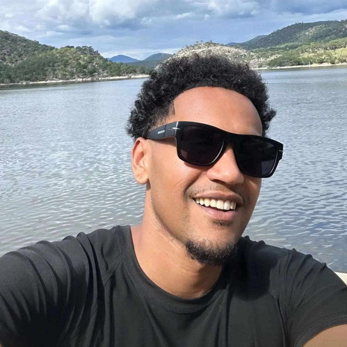 Hombre sonriente con gafas de sol cuadradas negras, montura de acetato y detalles metálicos frente a un lago.