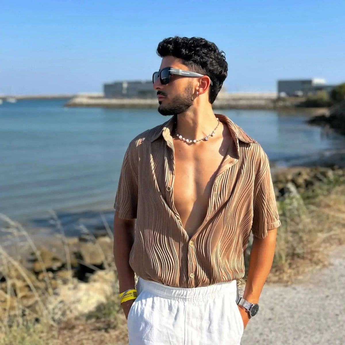 Hombre con gafas de sol cuadradas negras y detalle metálico, estilo veraniego con camisa beige plisada frente al mar.