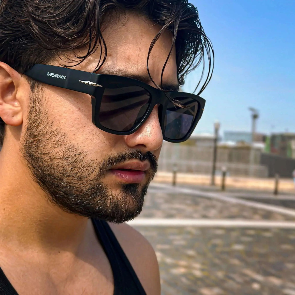 Hombre con barba usando gafas de sol negras cuadradas de estilo moderno con detalles metálicos en exteriores soleados.
