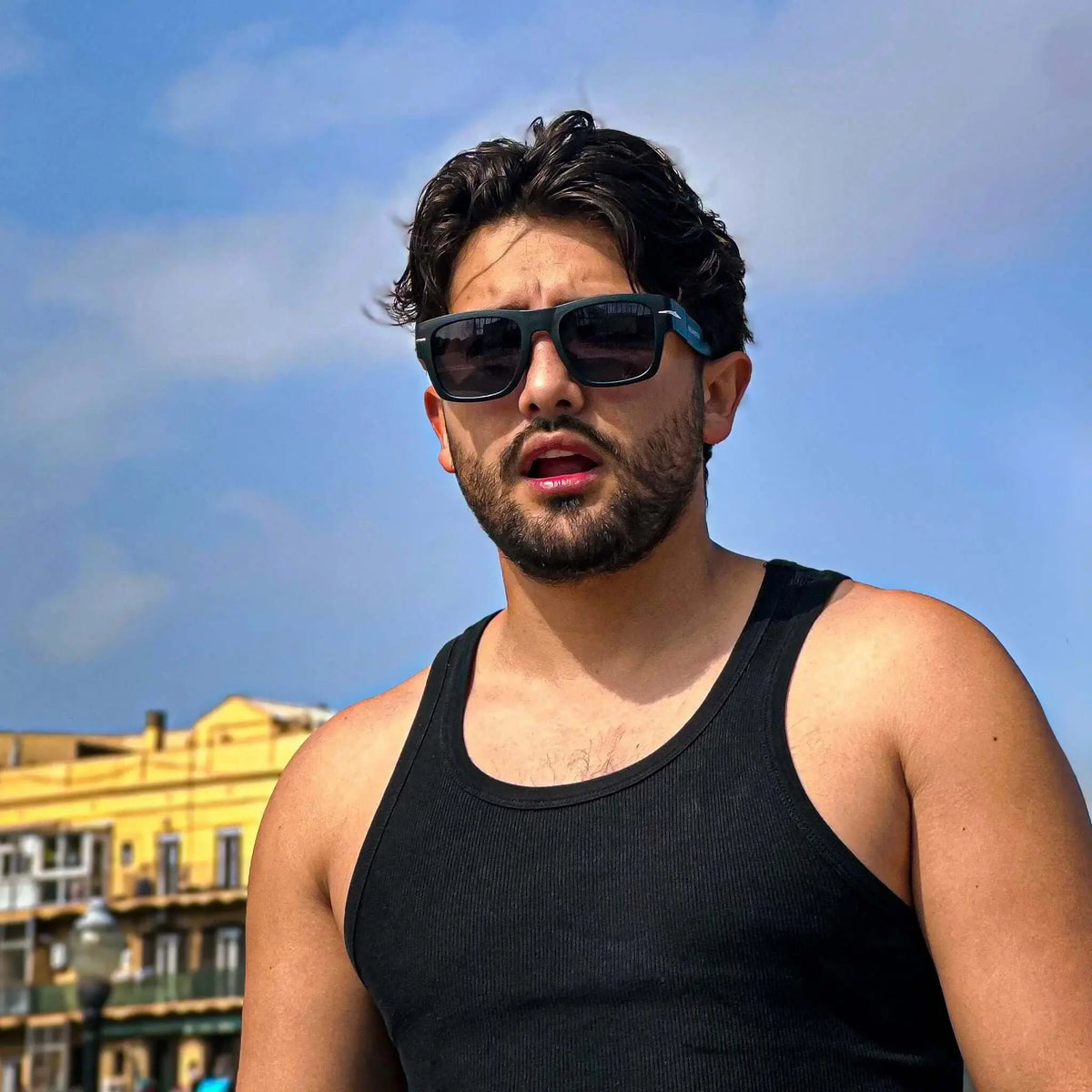 Hombre con gafas de sol cuadradas negras y detalle metálico, estilo casual con camiseta negra al aire libre.