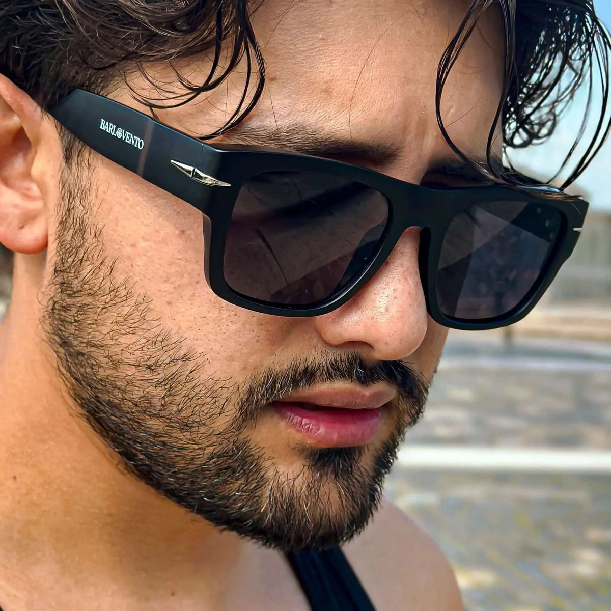 Hombre con barba usando gafas de sol negras cuadradas de estilo moderno con detalles metálicos y protección UV.