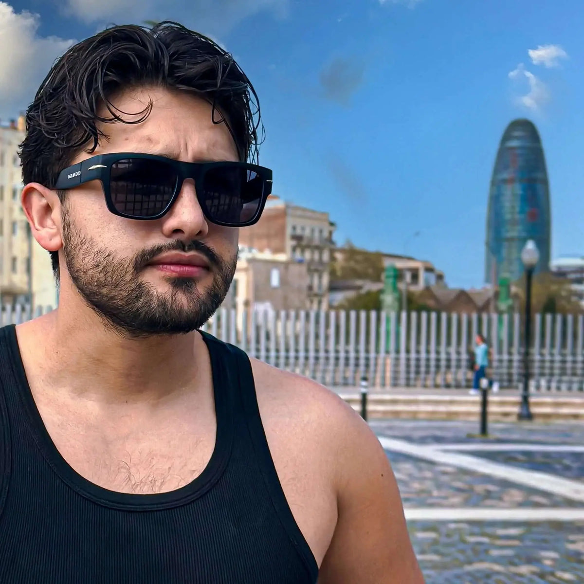 Hombre con gafas de sol cuadradas negras y detalle metálico, montura mate, estilo urbano frente a un edificio icónico.