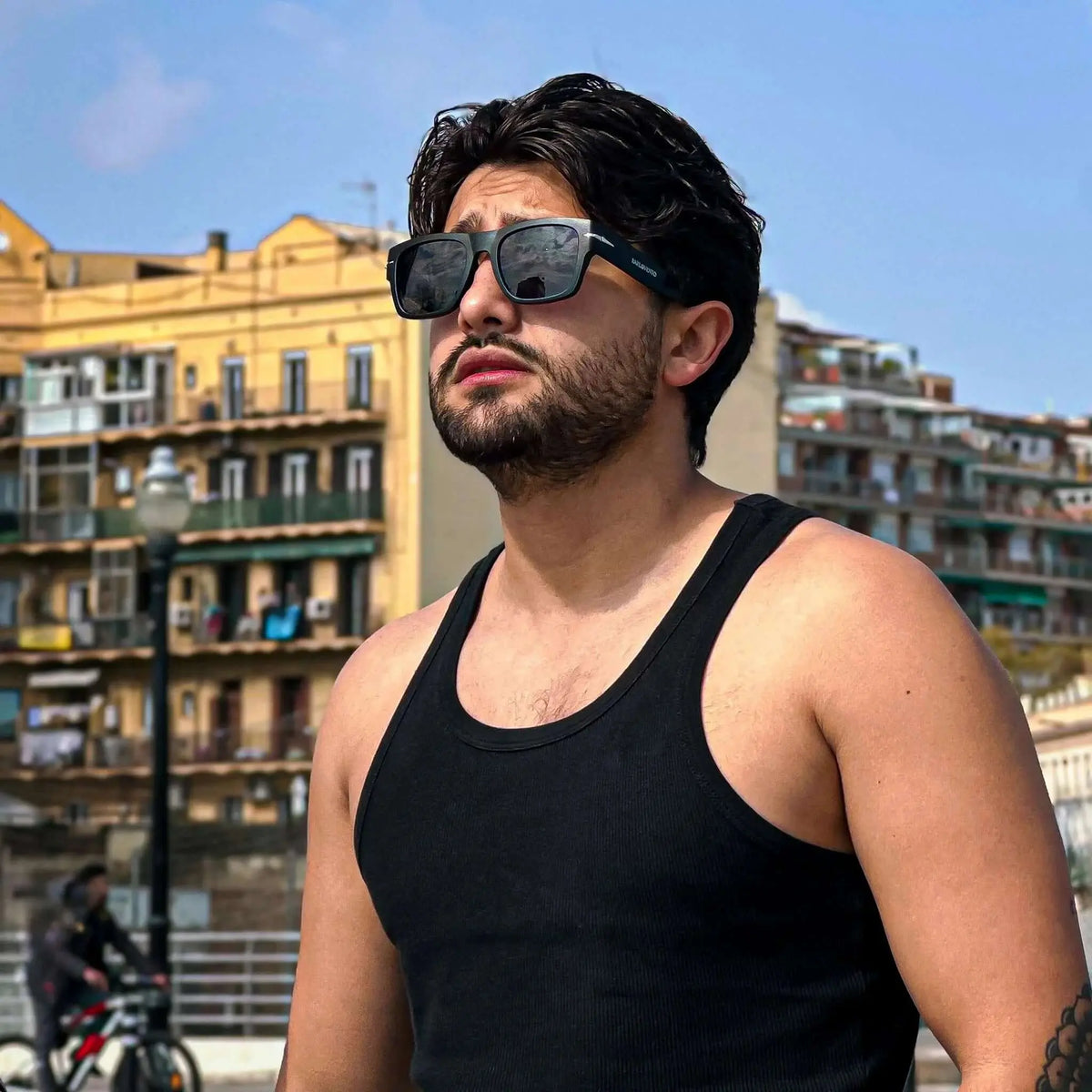 Hombre con gafas de sol cuadradas negras, montura de acetato con detalle metálico y protección UV en entorno urbano.