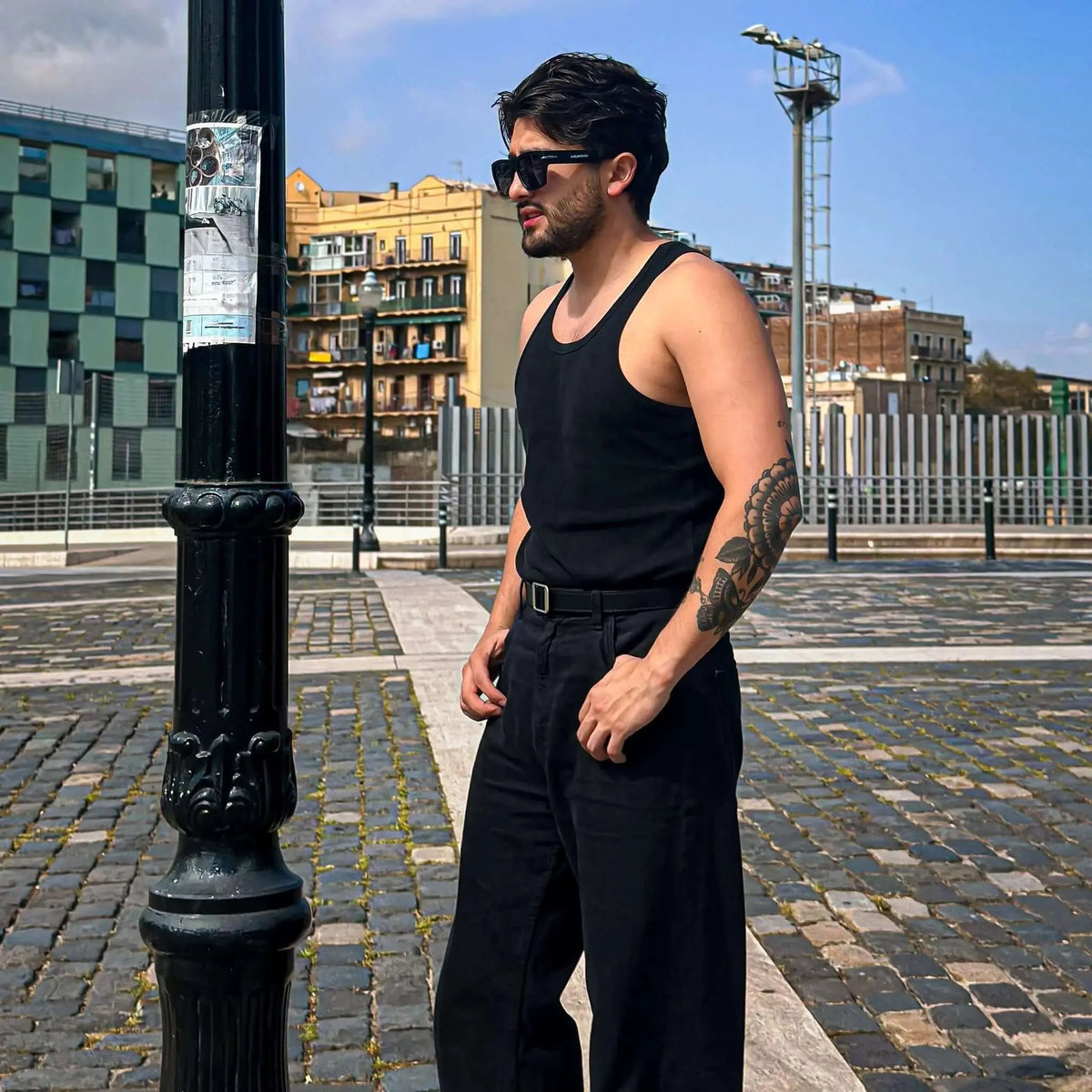 Hombre con gafas de sol cuadradas negras, montura con ribete metálico y protección UV, en entorno urbano casual.