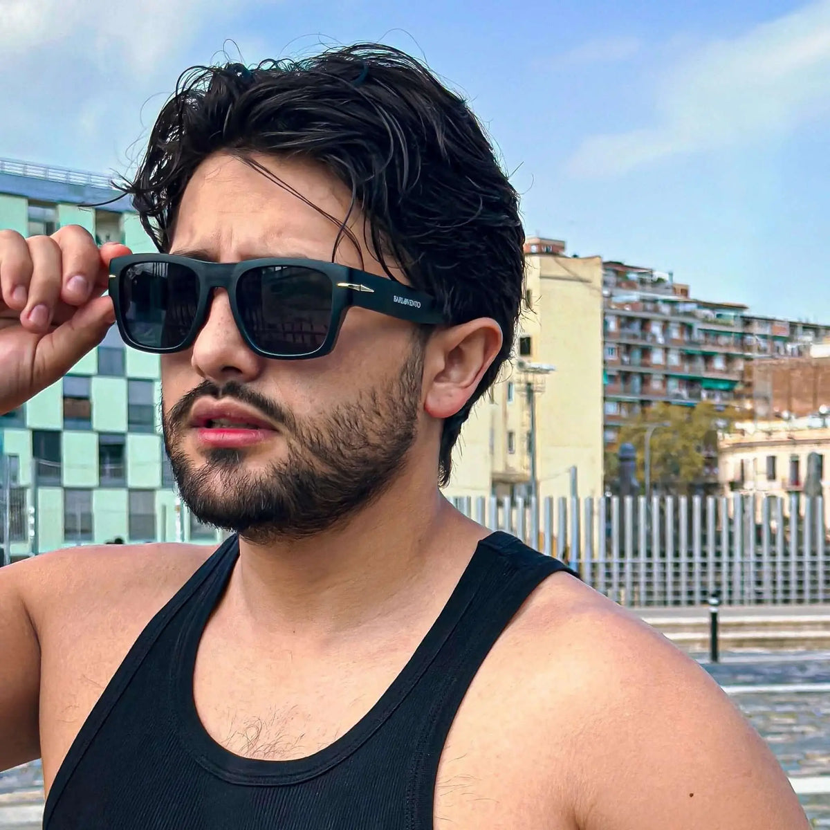 Hombre con barba y camiseta negra usando gafas de sol cuadradas negras con detalle metálico, estilo urbano y moderno.