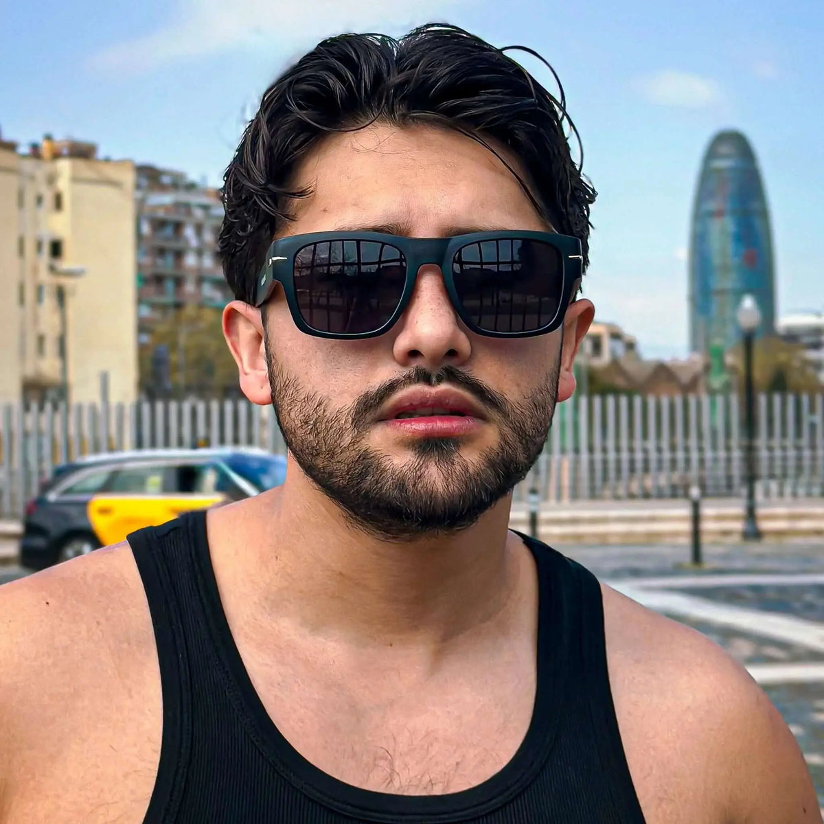 Hombre con camiseta negra y gafas de sol cuadradas negras con ribete metálico y protección UV en entorno urbano.