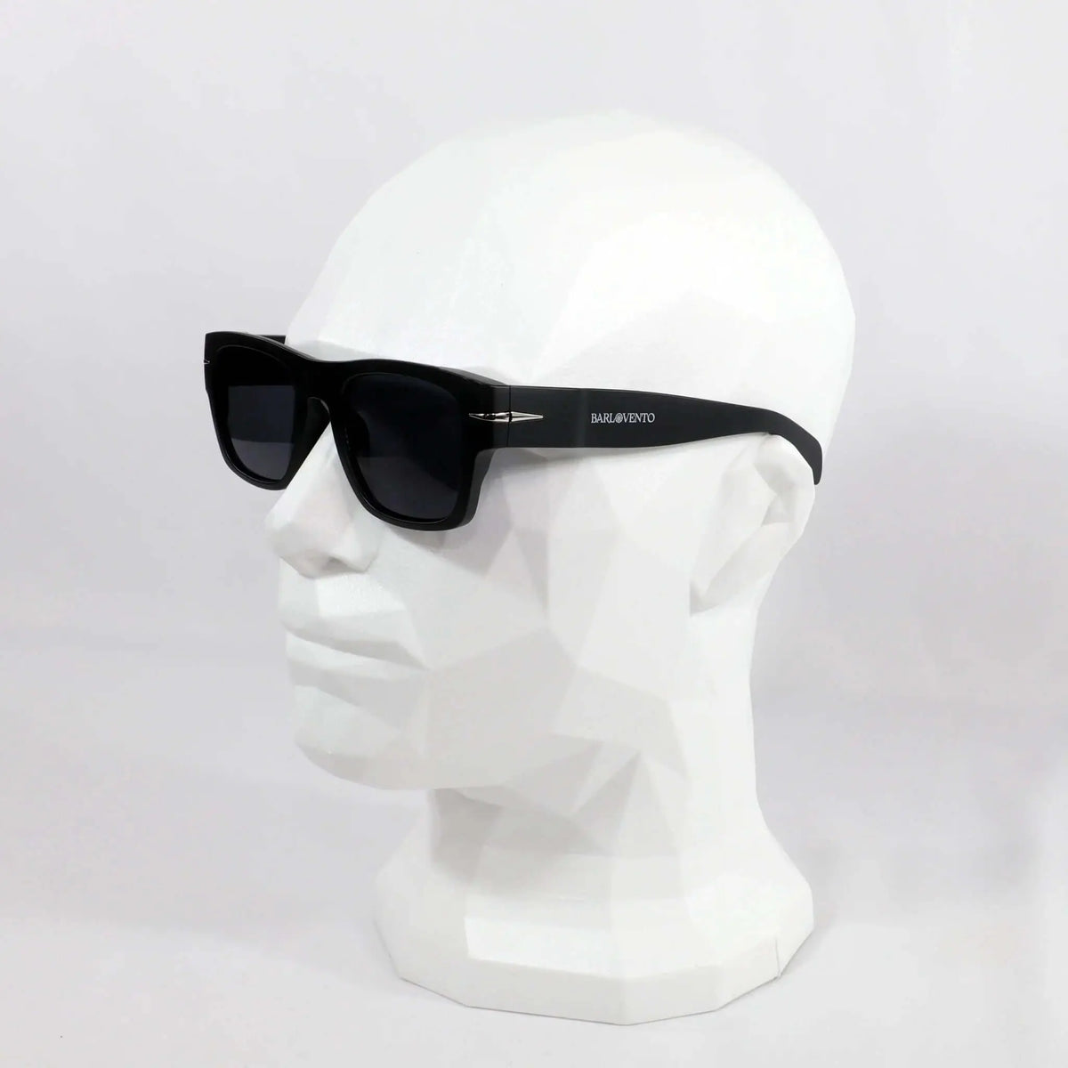 Gafas de sol negras cuadradas con montura gruesa, cristales oscuros y detalle metálico en maniquí blanco poligonal.