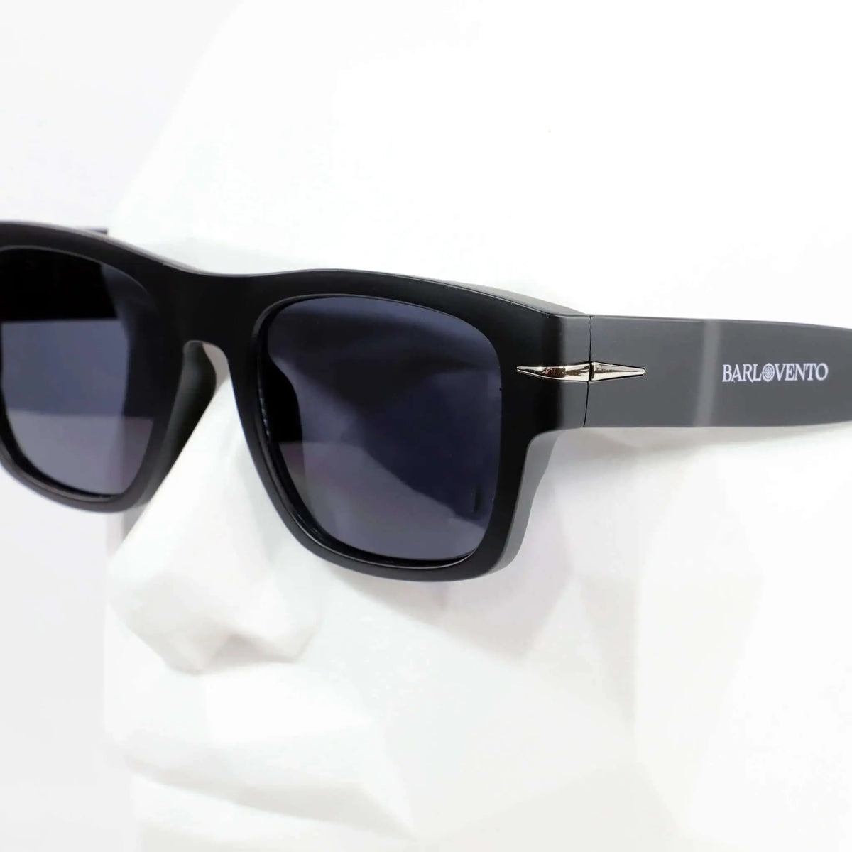Gafas de sol negras cuadradas con acabado mate, cristales oscuros y detalle metálico en la montura sobre maniquí.