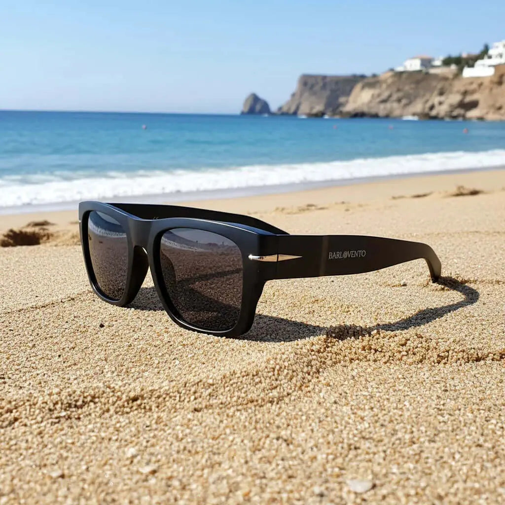 Gafas de sol negras cuadradas con detalle metálico, protección UV400, sobre la arena de una playa con mar de fondo.