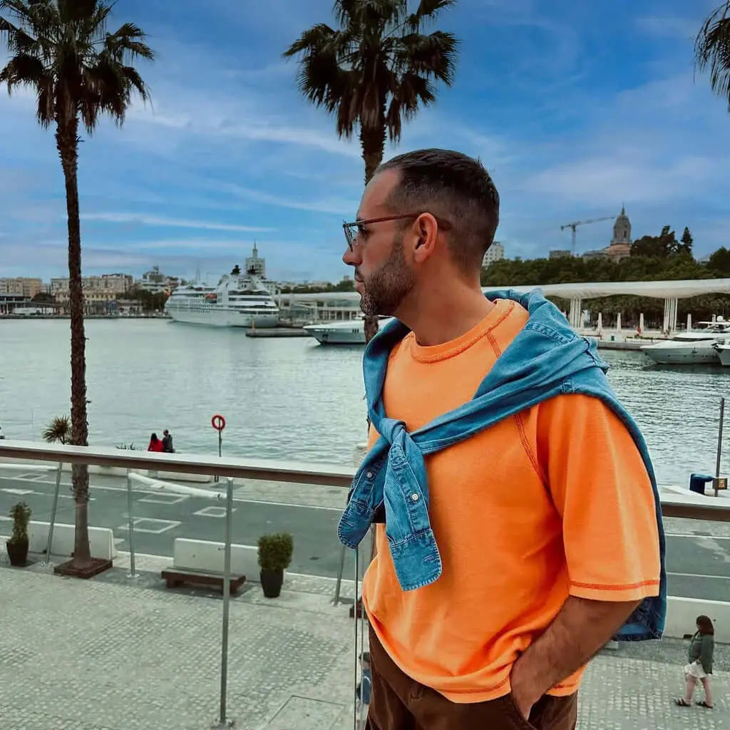 Hombre con gafas de sol de patillas de madera y lentes azules, vestido con camiseta naranja frente a un puerto marítimo.
