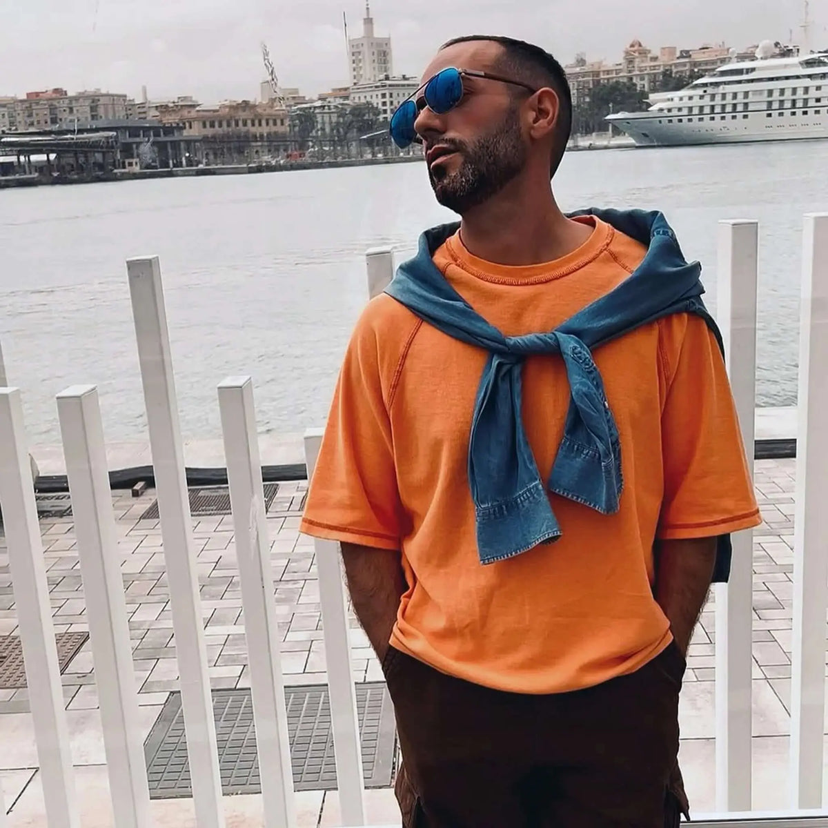 Hombre con gafas de sol de lentes azules y patillas de madera, estilo moderno frente al puerto.