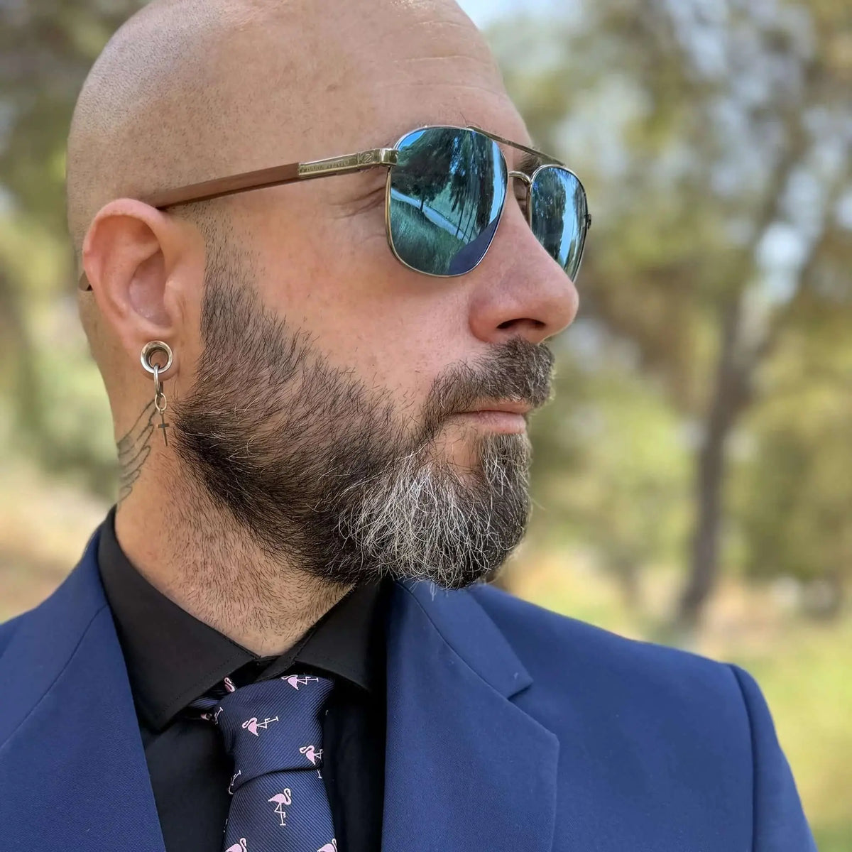 Hombre con gafas de sol tipo aviador, lentes azules y patillas de madera, vistiendo traje azul y camisa negra.
