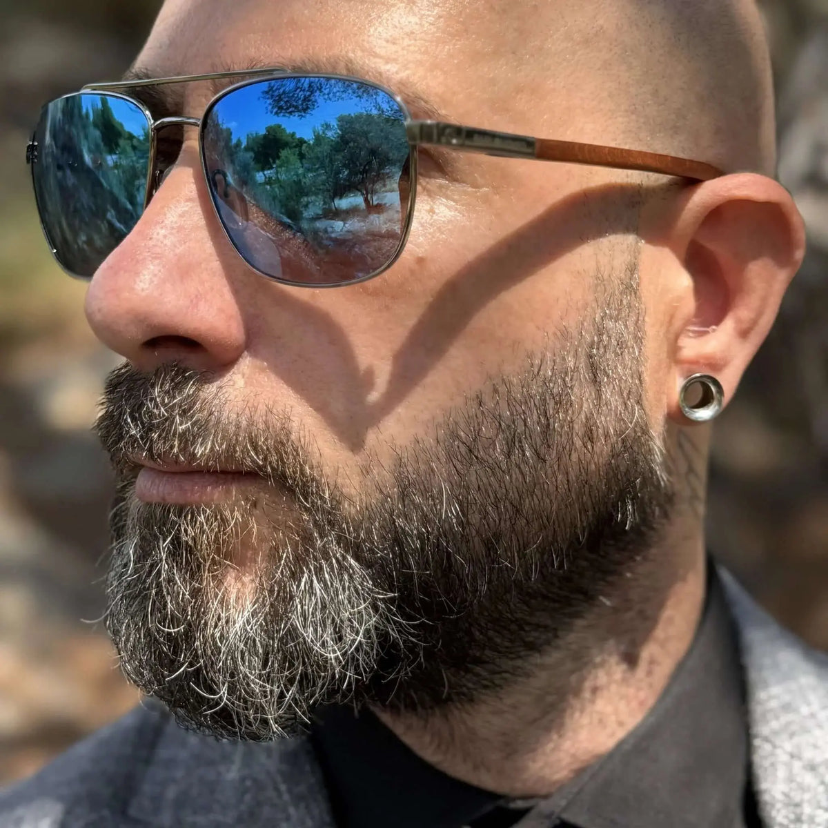Hombre barbudo con gafas de sol tipo aviador, lentes azules espejadas y patillas de madera en entorno exterior.