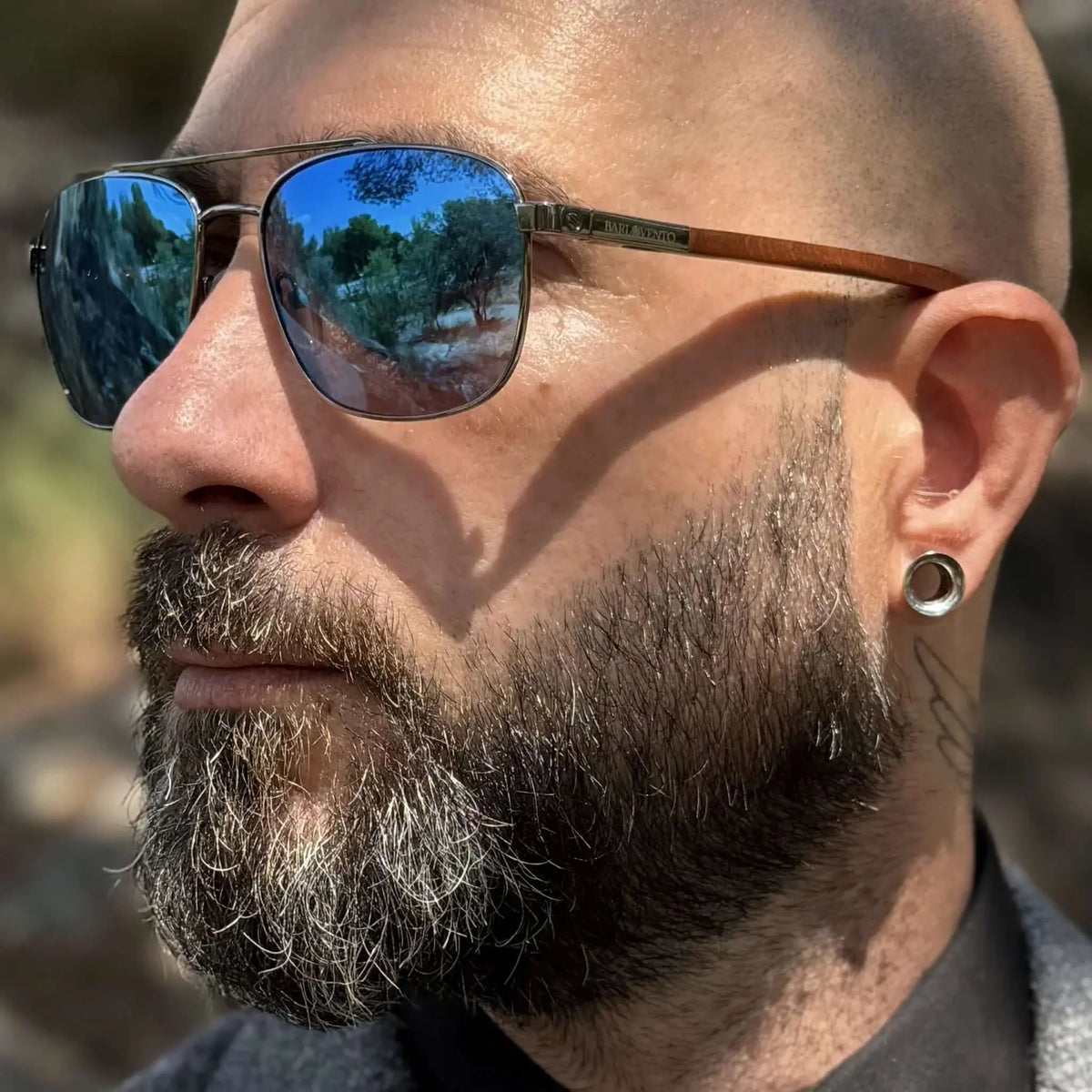 Gafas de sol estilo aviador con lentes espejo azules y patillas de madera, puestas en un hombre con barba y dilatación.