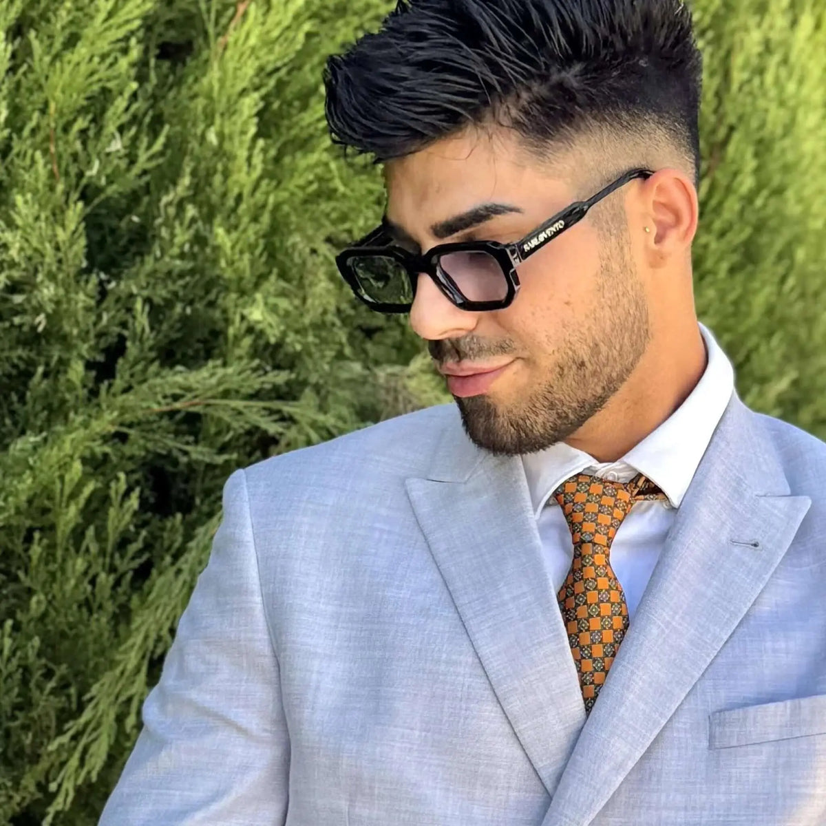 Gafas de sol negras de estilo rectangular con lentes azuladas, usadas por un hombre con traje gris y corbata estampada.