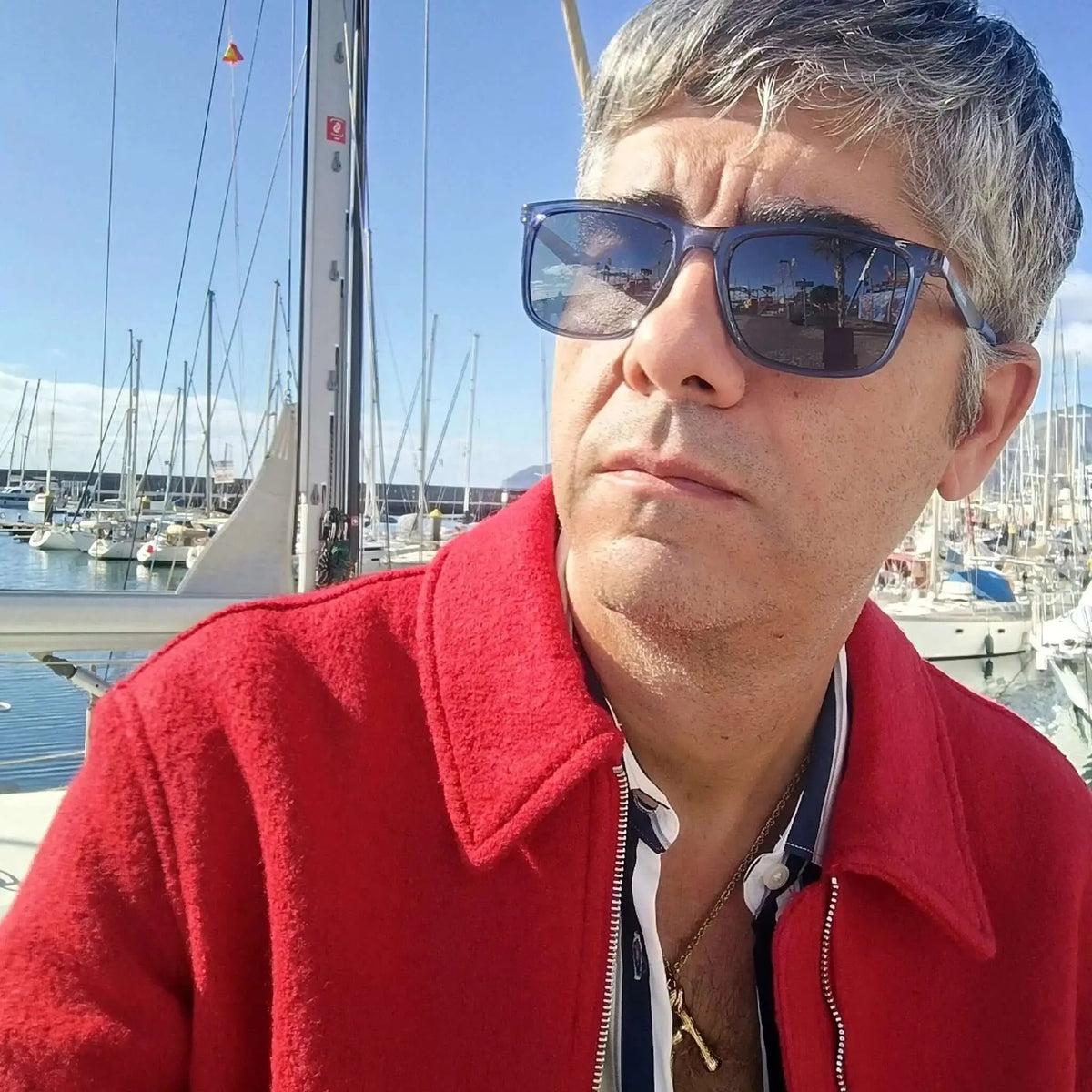 Hombre con chaqueta roja y gafas de sol de montura azul traslúcida estilo wayfarer, posando en un puerto con veleros.