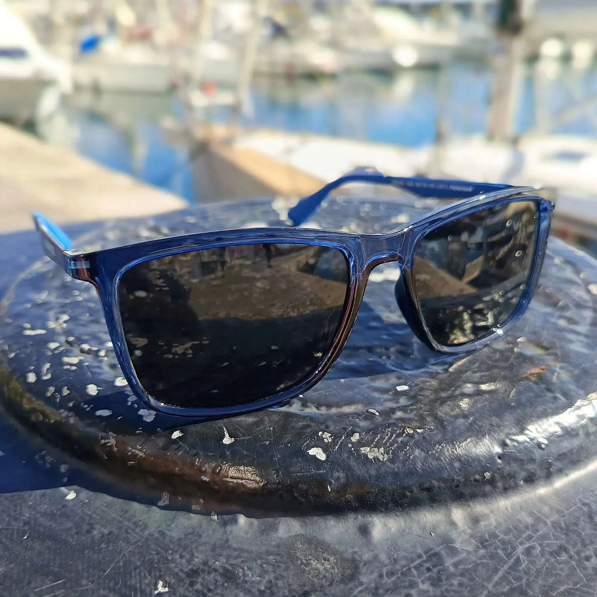 Gafas de sol de montura azul translúcida y lentes oscuras sobre superficie náutica con fondo de puerto desenfocado.