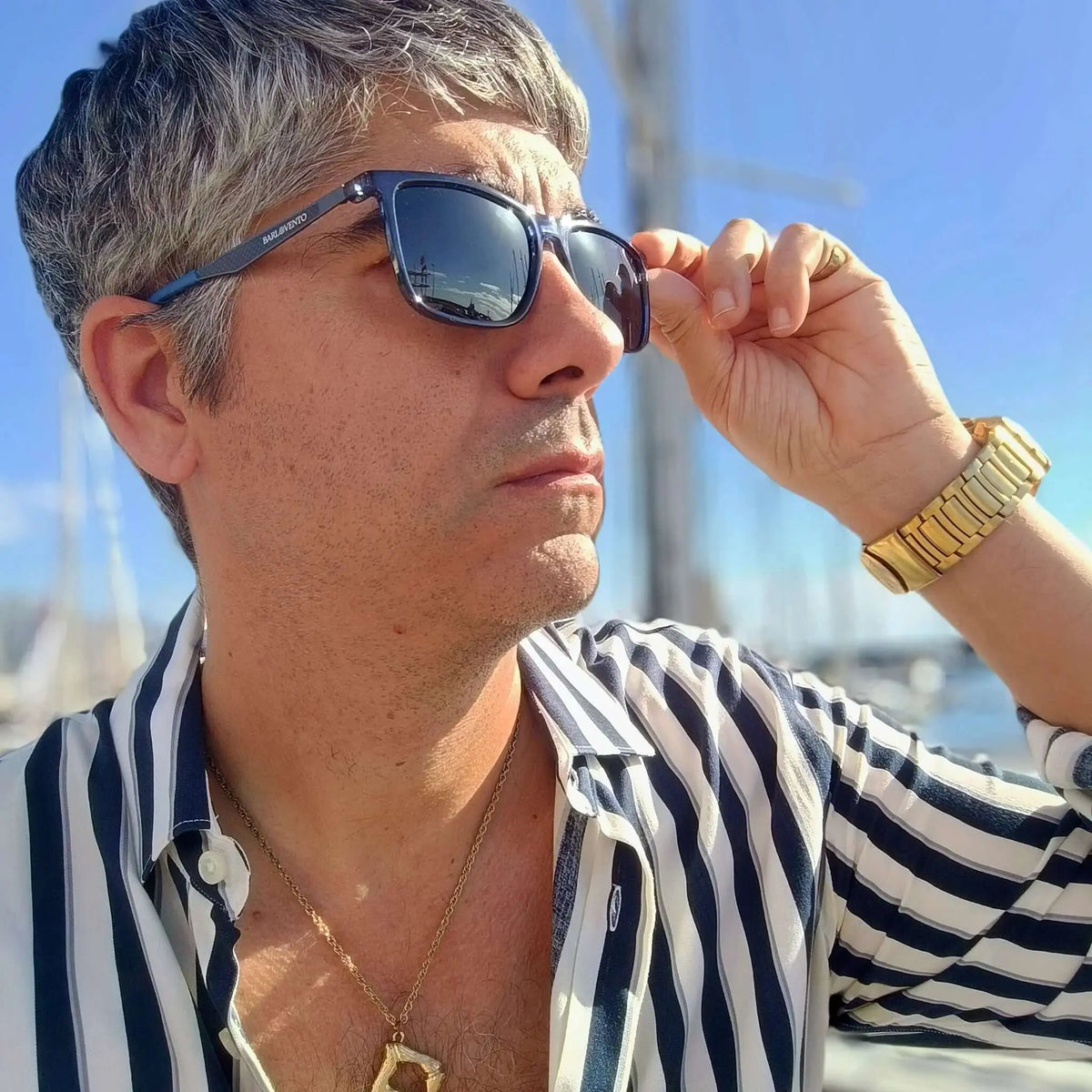 Hombre con camisa a rayas y gafas de sol de montura azul cuadrada, estilo náutico, con cristales oscuros reflejantes.