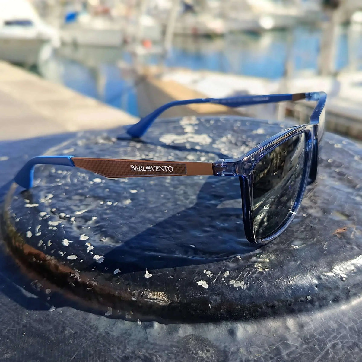 Gafas de sol de montura azul traslúcida y patillas metálicas color cobre sobre superficie oscura frente a un puerto.