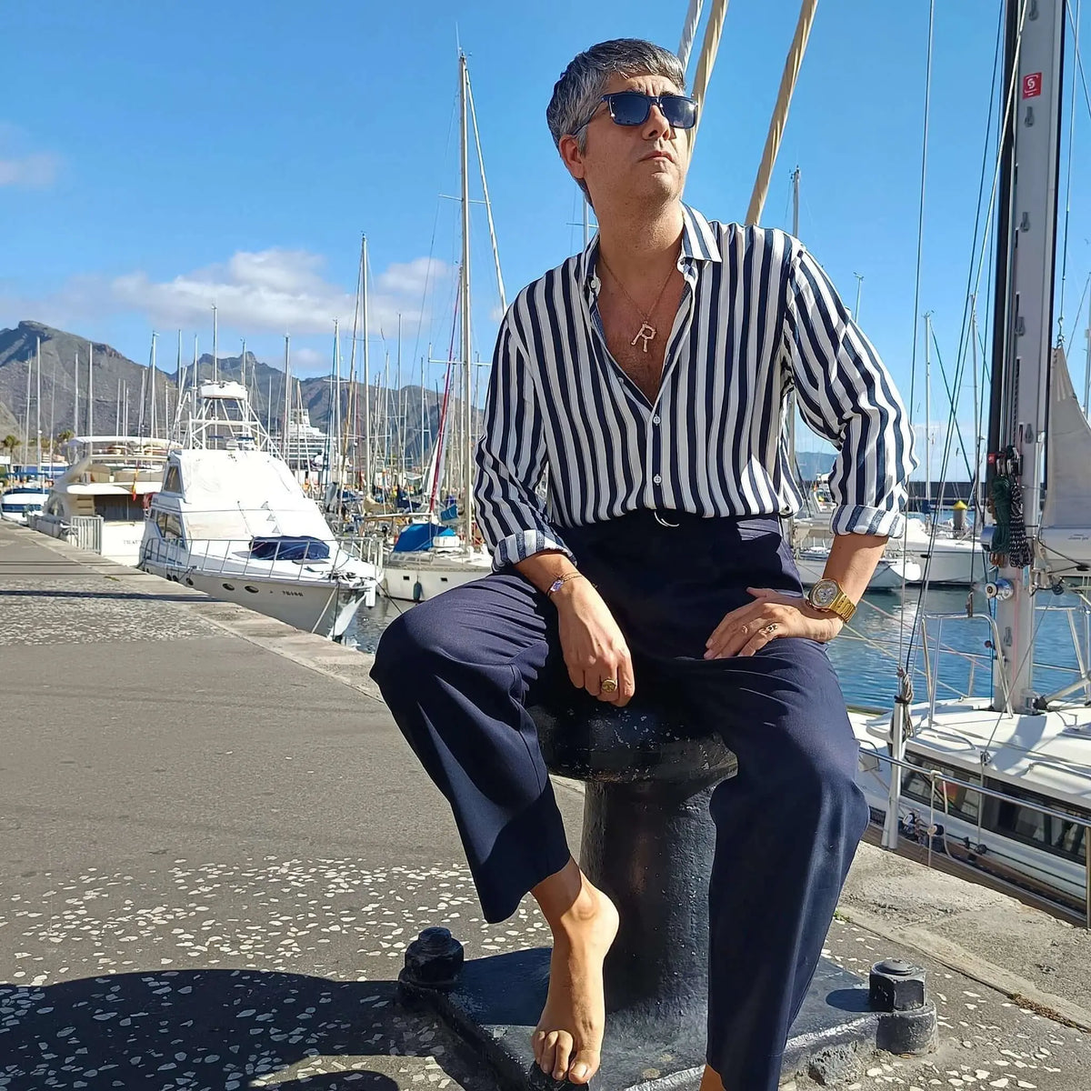 Hombre con gafas de sol de montura azul y lentes oscuras, sentado en un puerto deportivo con barcos al fondo.